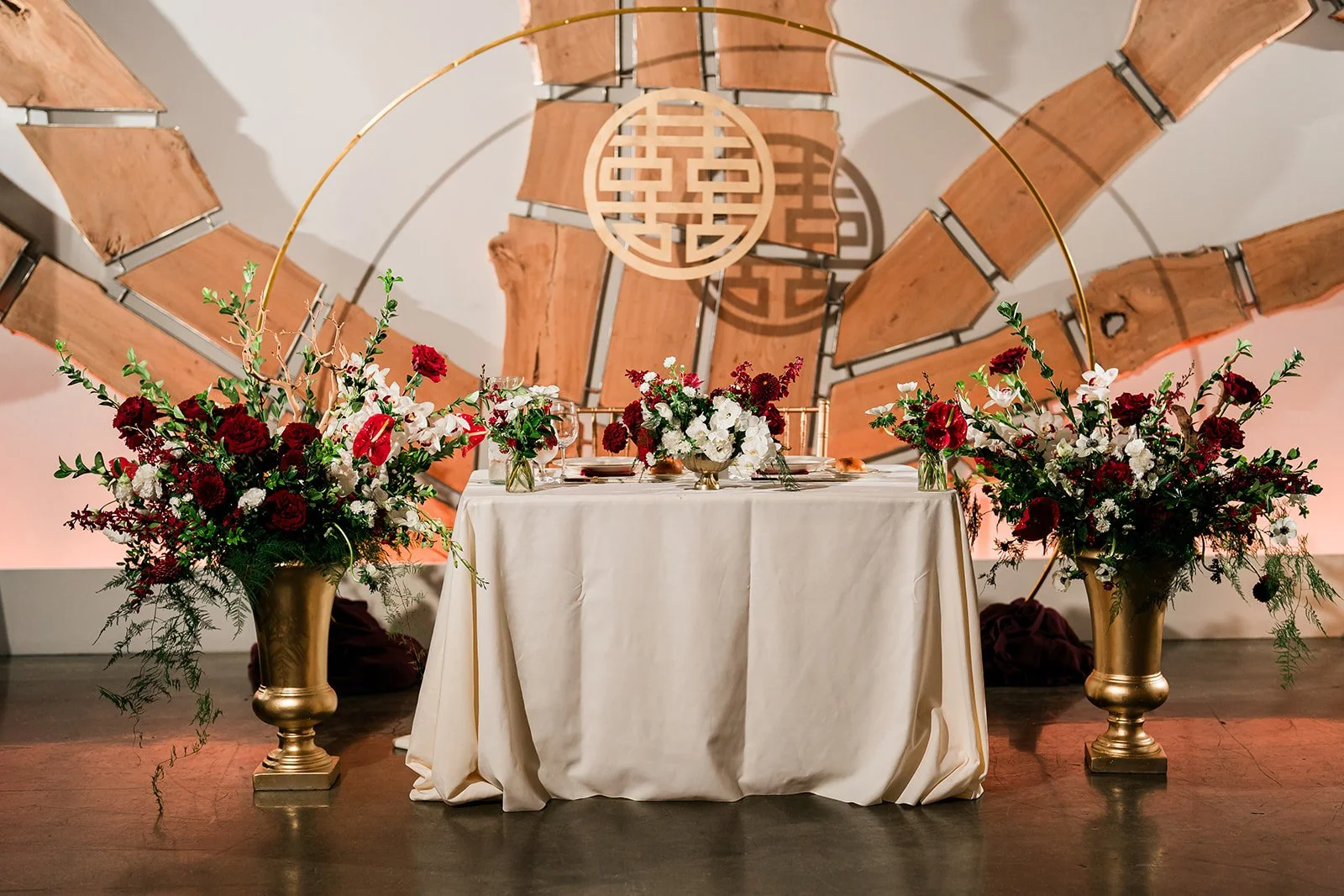 Sweetheart table with white tablecloth adorned with floral arrangements in gold vases, set against a Double Happiness sign with circular design in front of the signature tree at The Pearl SF.