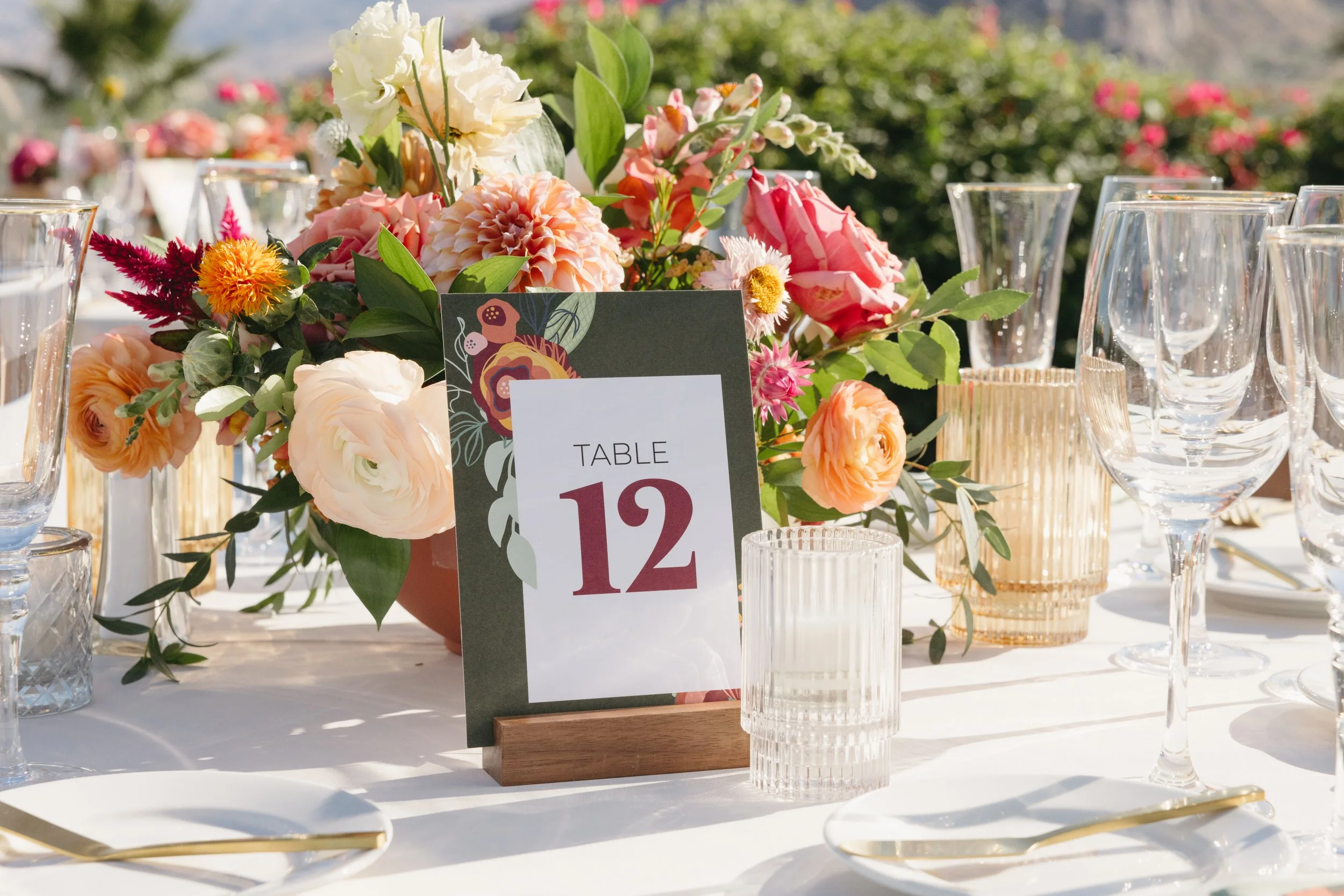 Elegant table setting with a floral centerpiece, clear glassware, gold utensils, and a table number sign indicating "Table 12" at an outdoor event.
