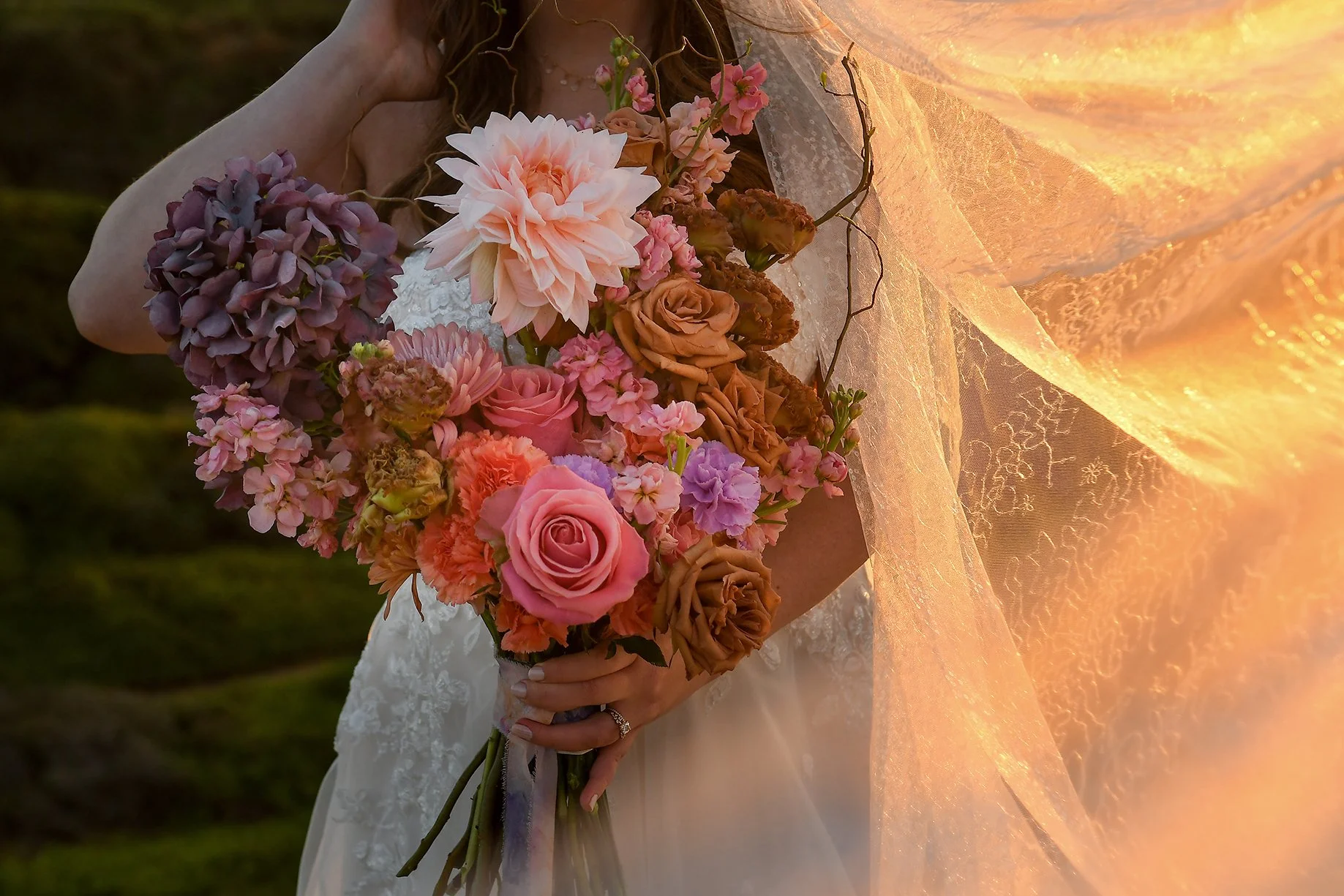 A bride holding a large bouquet of pink, purple, cream, and brown flowers, standing outdoors with a glowing veil with lace details reflecting sunlight.