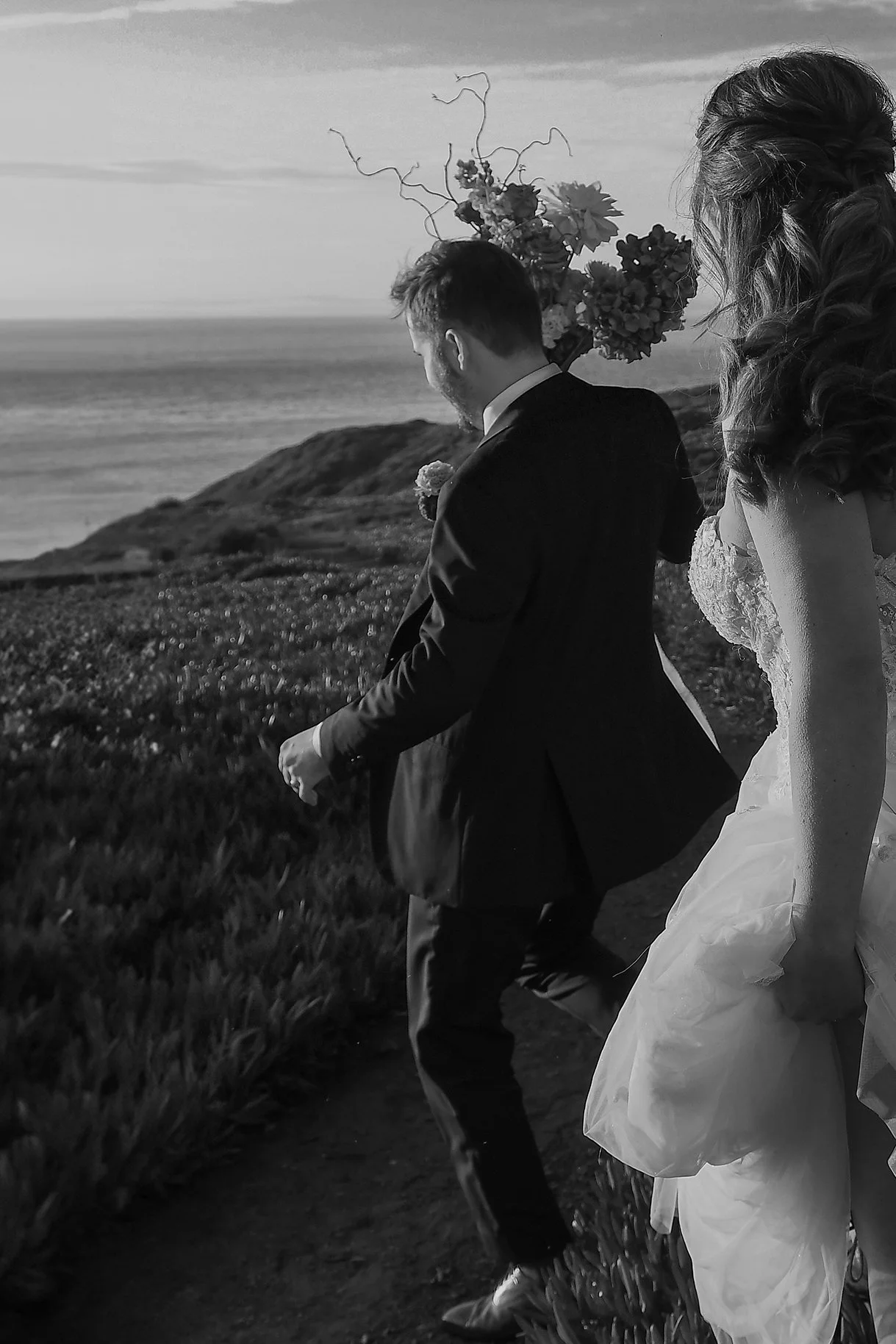 Black and white photo of a bride and groom walking along a coastal path during sunset, with the ocean in the background.
