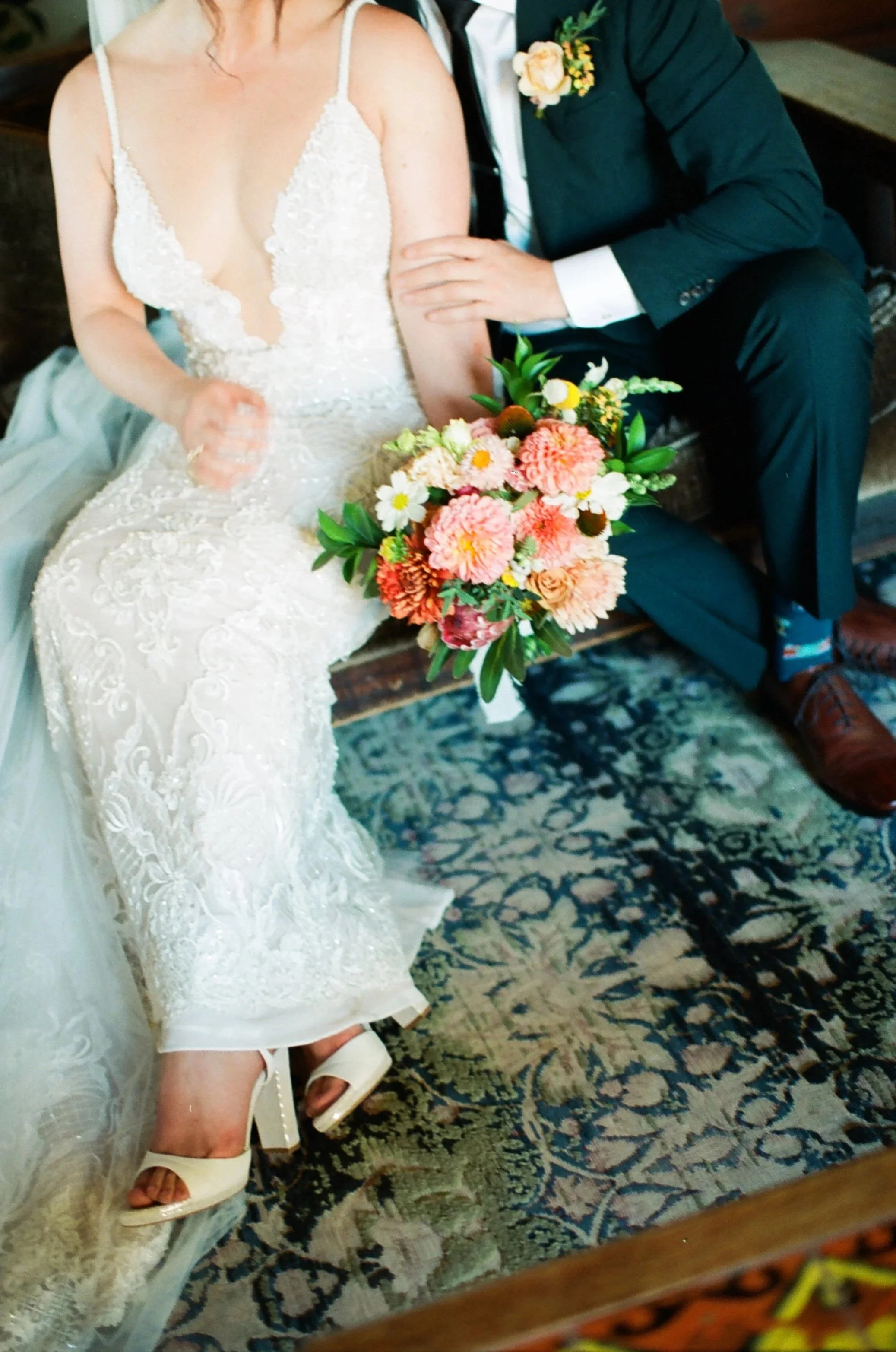 A bride in a lace wedding dress and heels sitting beside a groom in a black suit holding a bouquet of pink, orange, white, and yellow flowers, both sitting on a wooden bench with a patterned rug underneath.