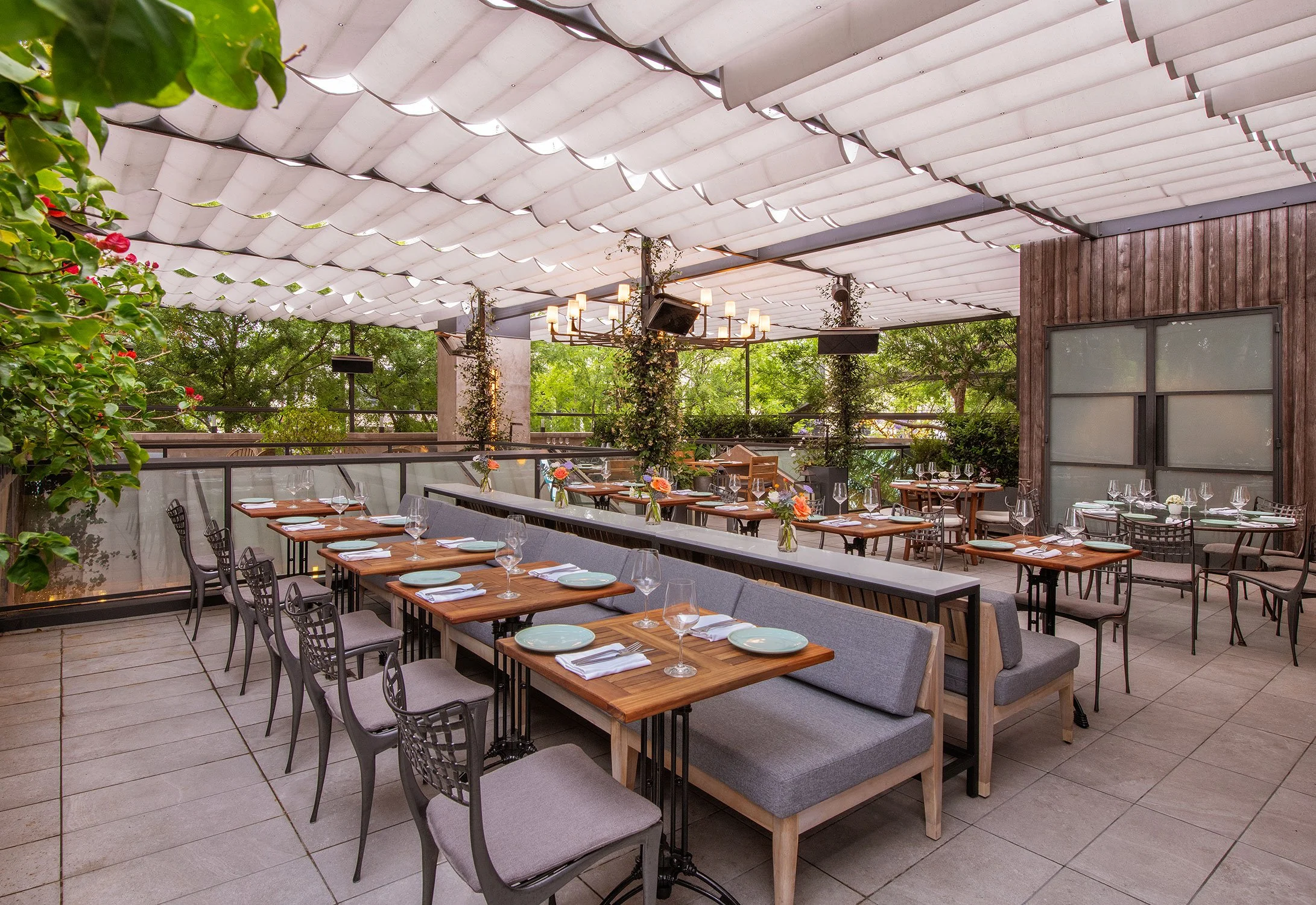 A restaurant patio with wooden tables and metal chairs, decorated with napkins and glassware, under a retractable white canopy, surrounded by greenery and string lights.