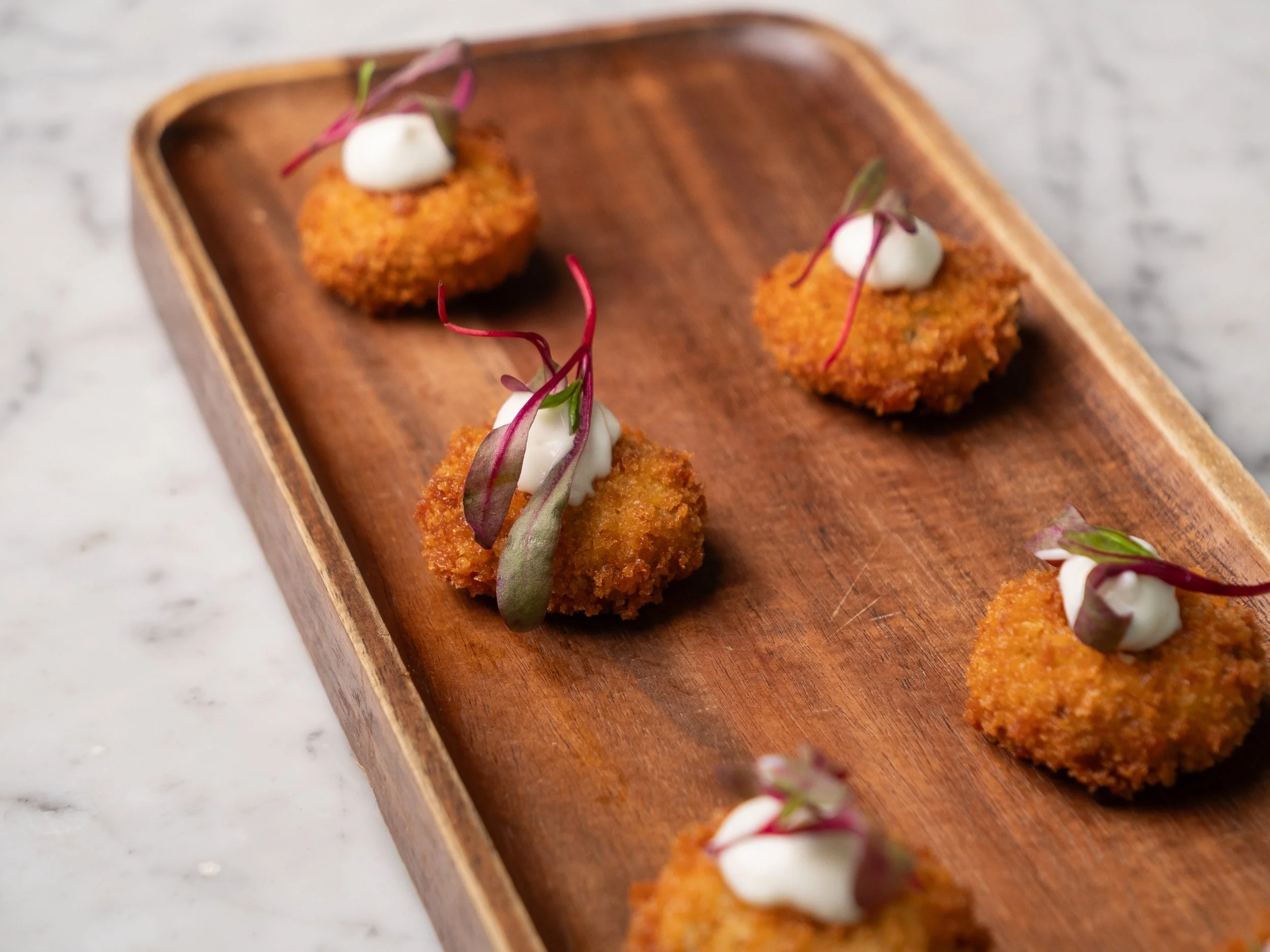 Breaded and fried bite-sized appetizer pieces topped with dollops of white sauce and garnished with microgreens, served on a wooden tray.