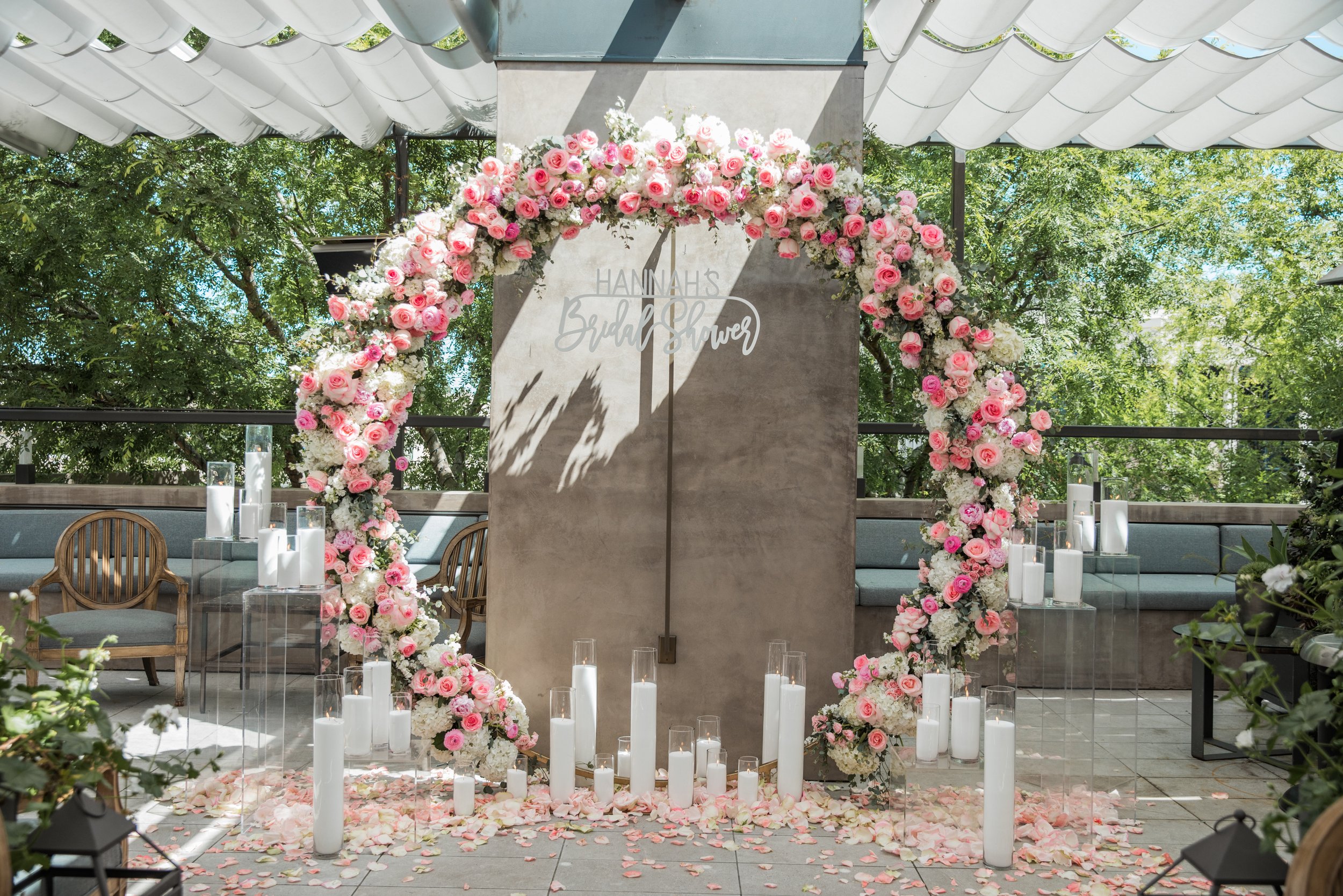 A wedding alter decorated with pink and white roses and white candles on the floor, set outdoors with trees in the background.