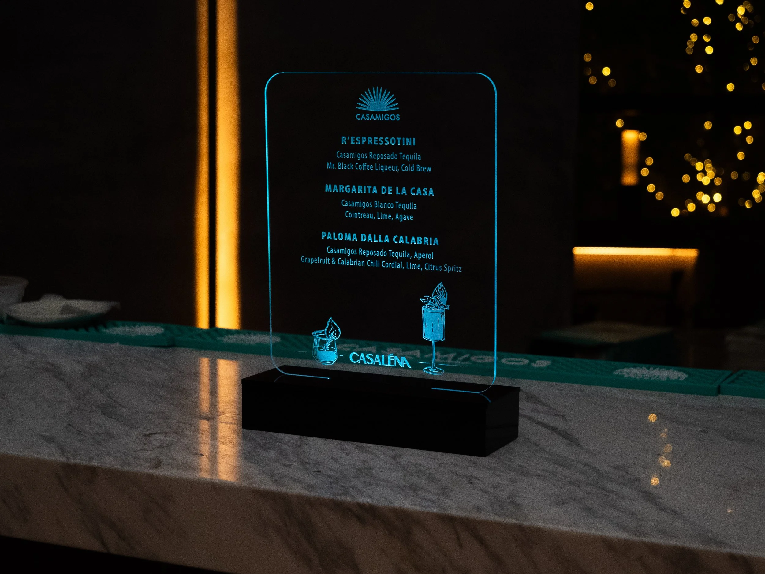 A clear acrylic menu stand displaying a drink menu with blue text, placed on a marble countertop in a dimly lit setting, with blurred yellow lights in the background.