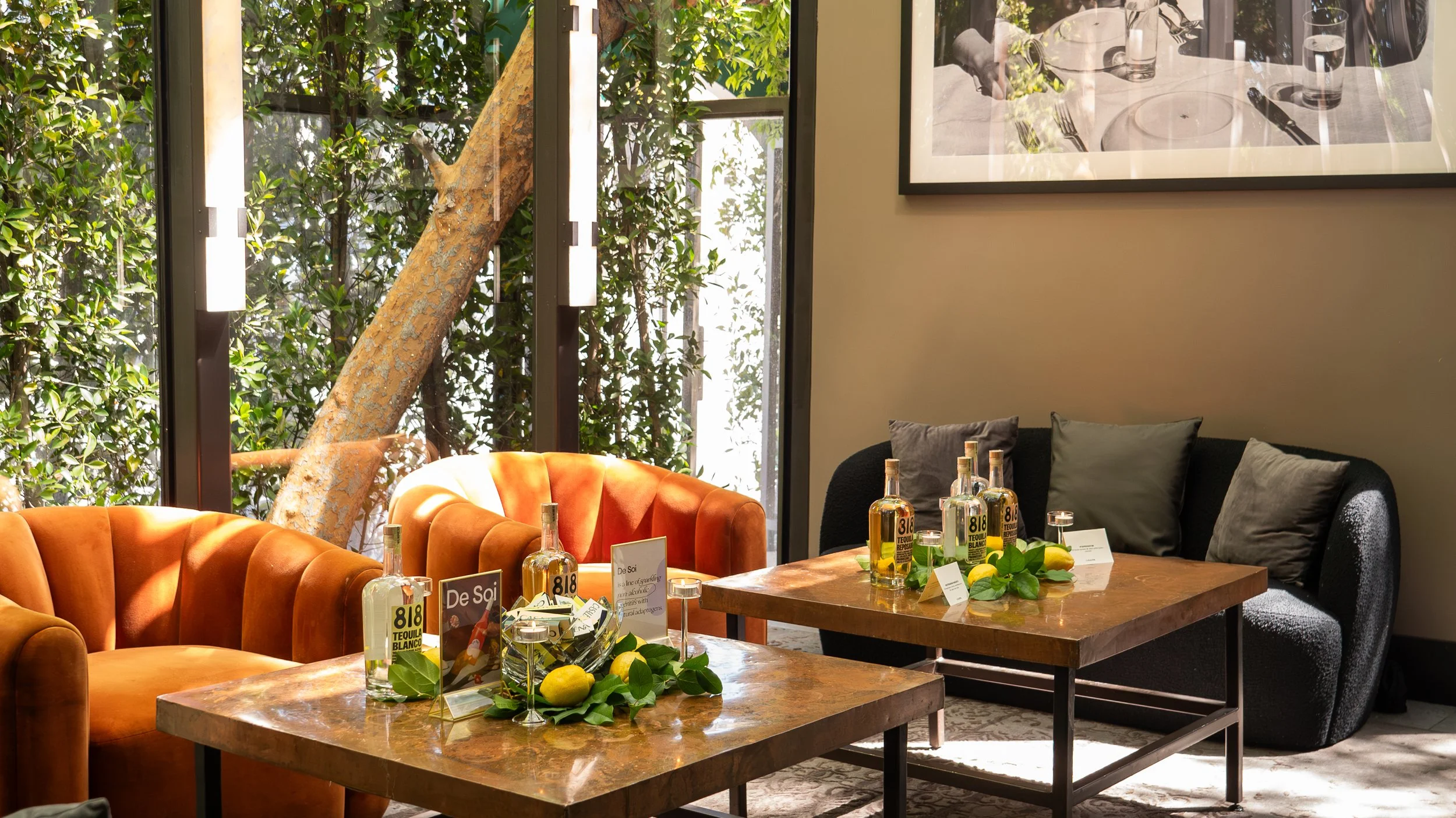 Indoor setting with tables and seating, including a tan velvet sofa and a dark upholstered chair, decorated with bottles of tequila, lime wedges, and small signs, with large windows, greenery outside, and a framed black and white photograph on the wa