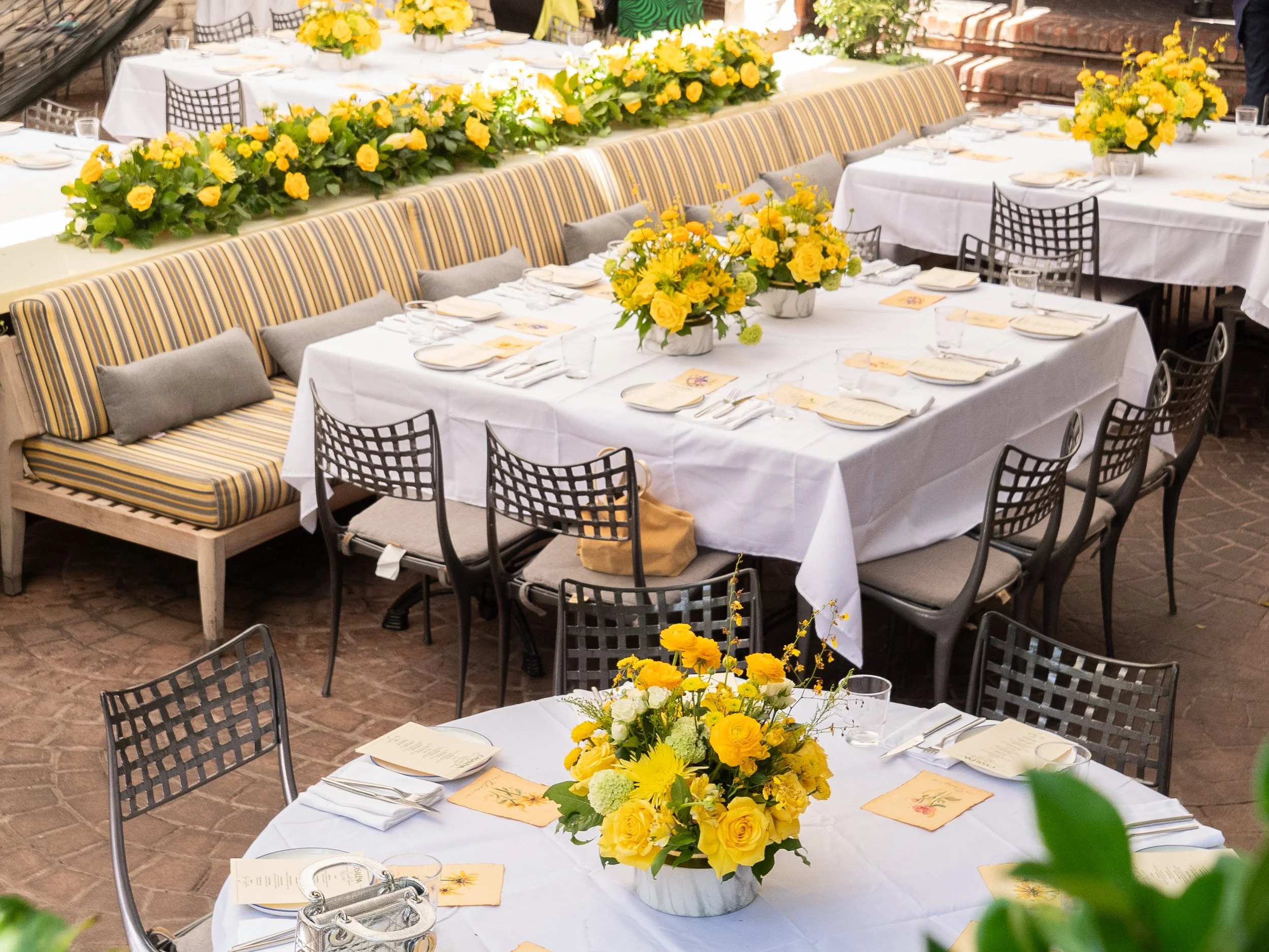 A banquet table decorated with vibrant yellow and white floral arrangements, white tablecloths, and place settings, set for a celebration or event in a rustic indoor space with exposed brick walls and brick flooring.