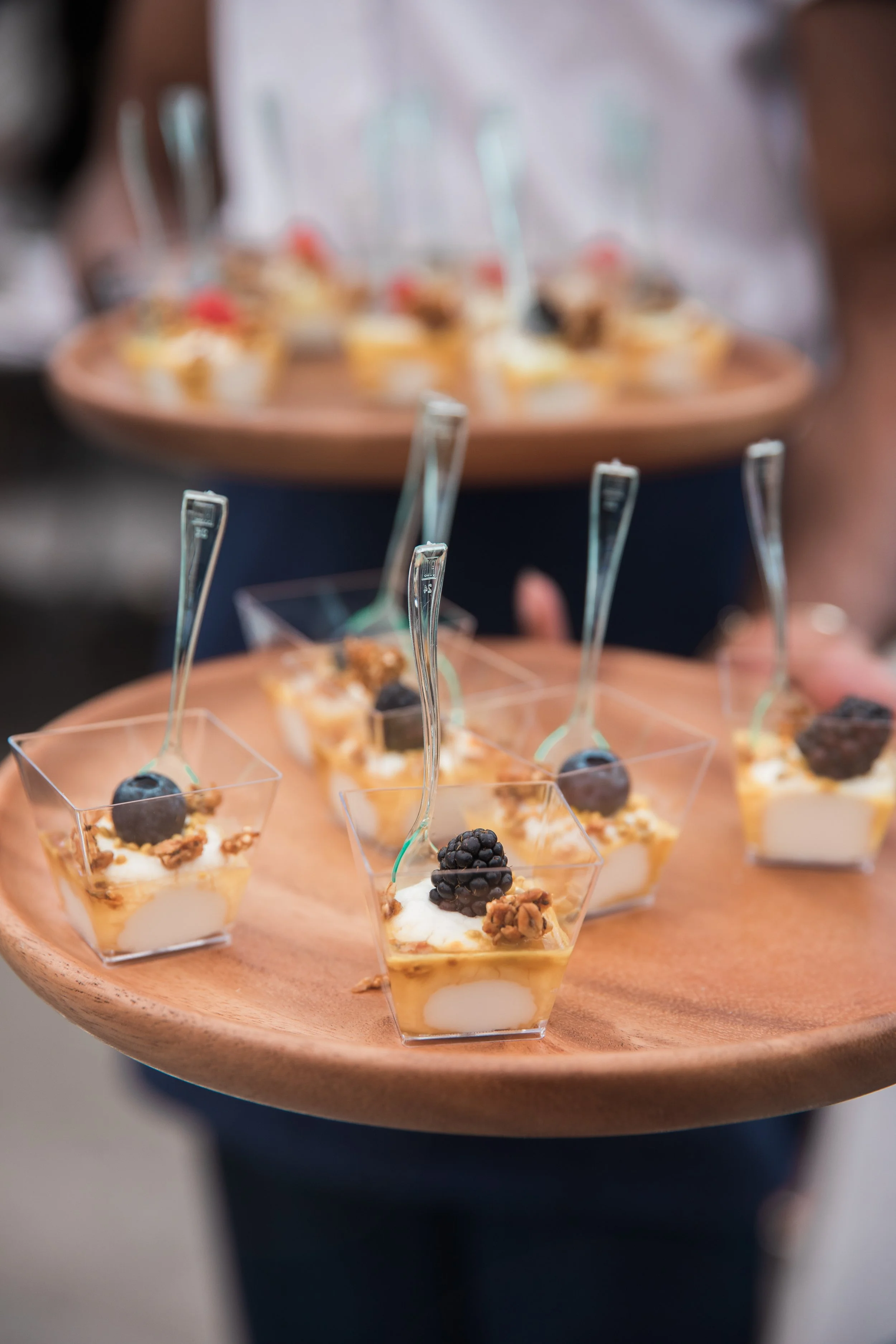 Close-up of a wooden serving tray holding small desserts in clear cups, topped with blackberries, walnuts, and other toppings, with a blurred background.