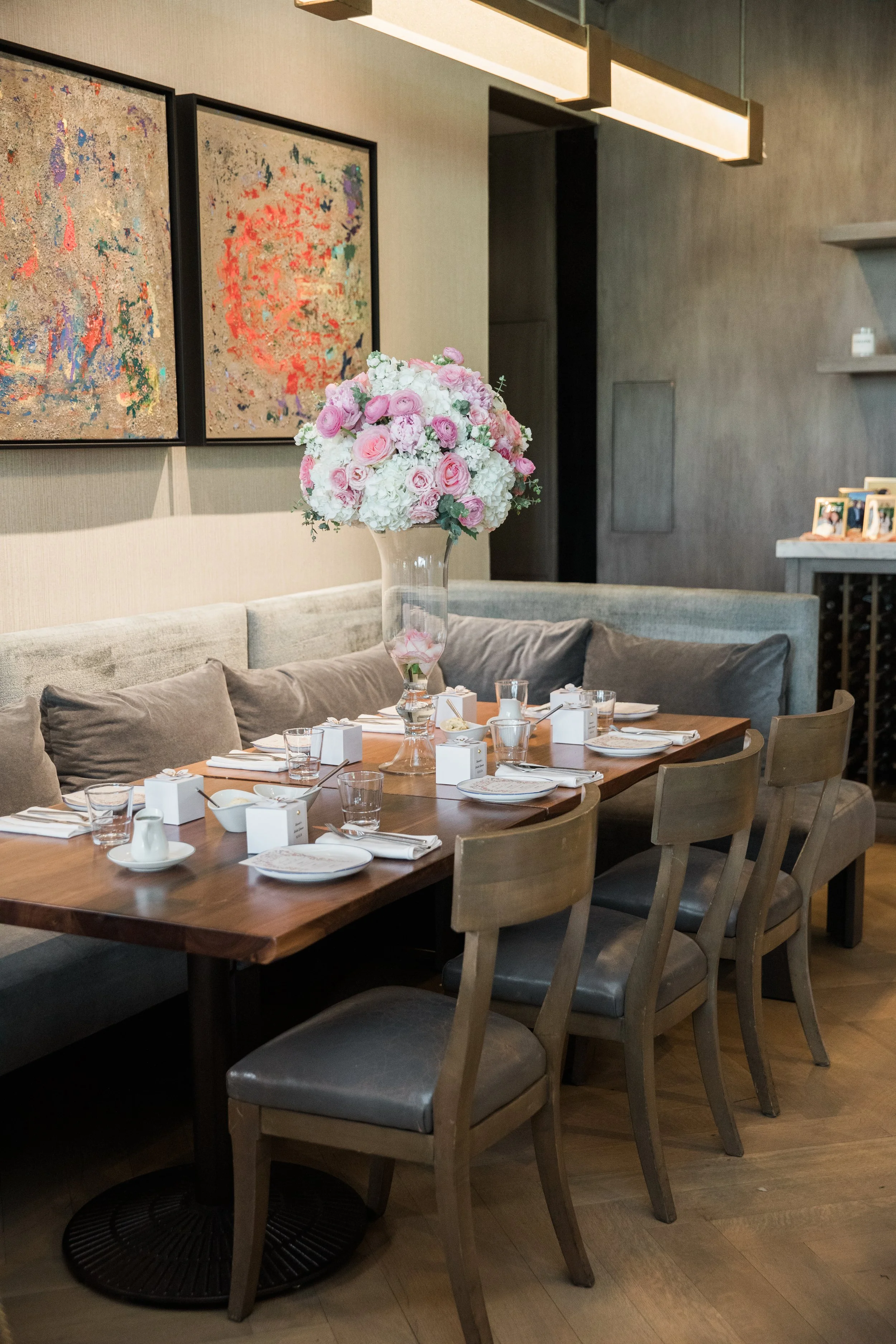 A dining table set for a meal with a large floral centerpiece in a glass vase, surrounded by chairs and small boxes, in a modern dining area with framed artwork on gray walls and a contemporary light fixture overhead.