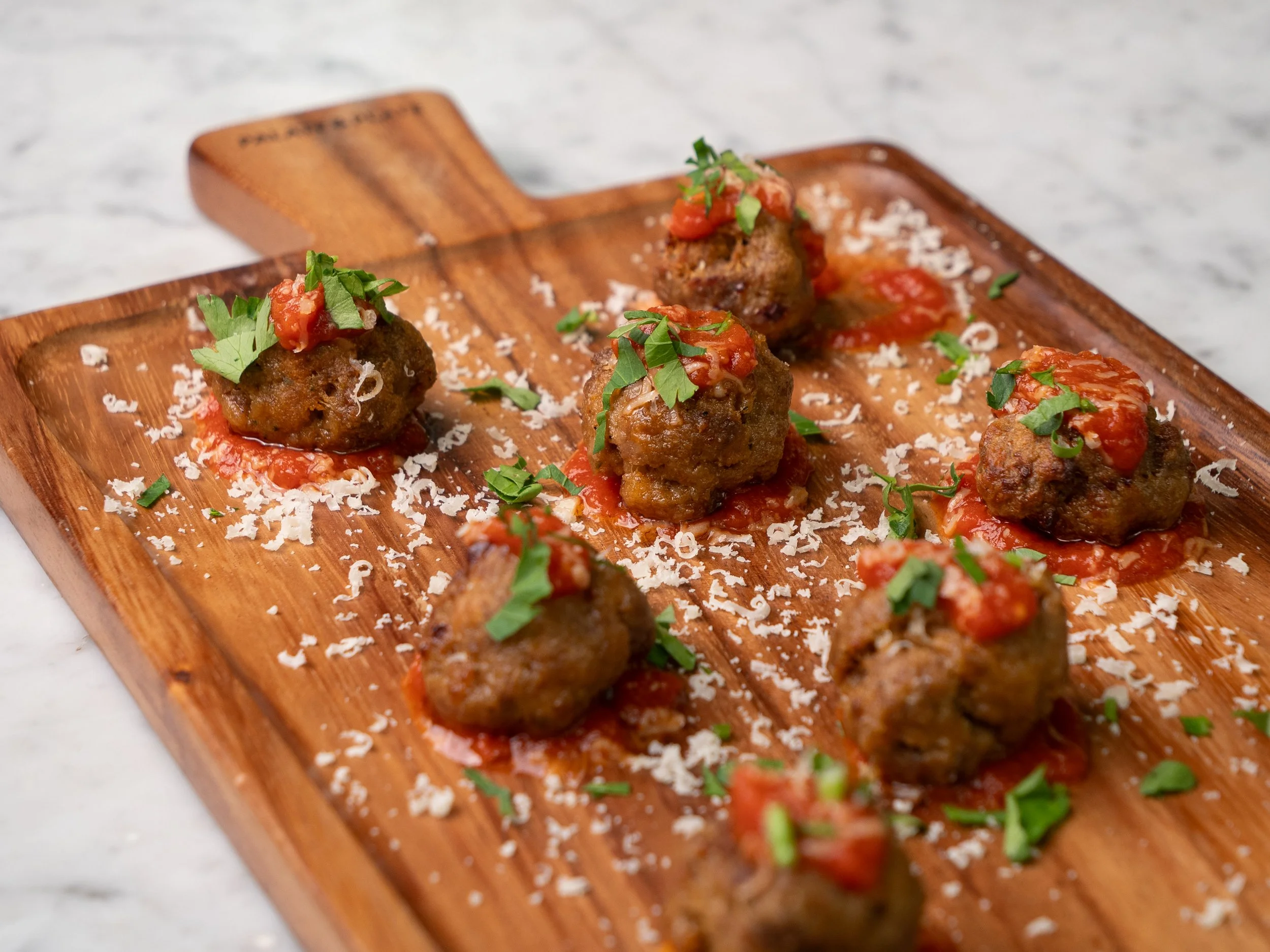 Close-up of a wooden serving board with several meatballs topped with tomato sauce, chopped herbs, and grated cheese.