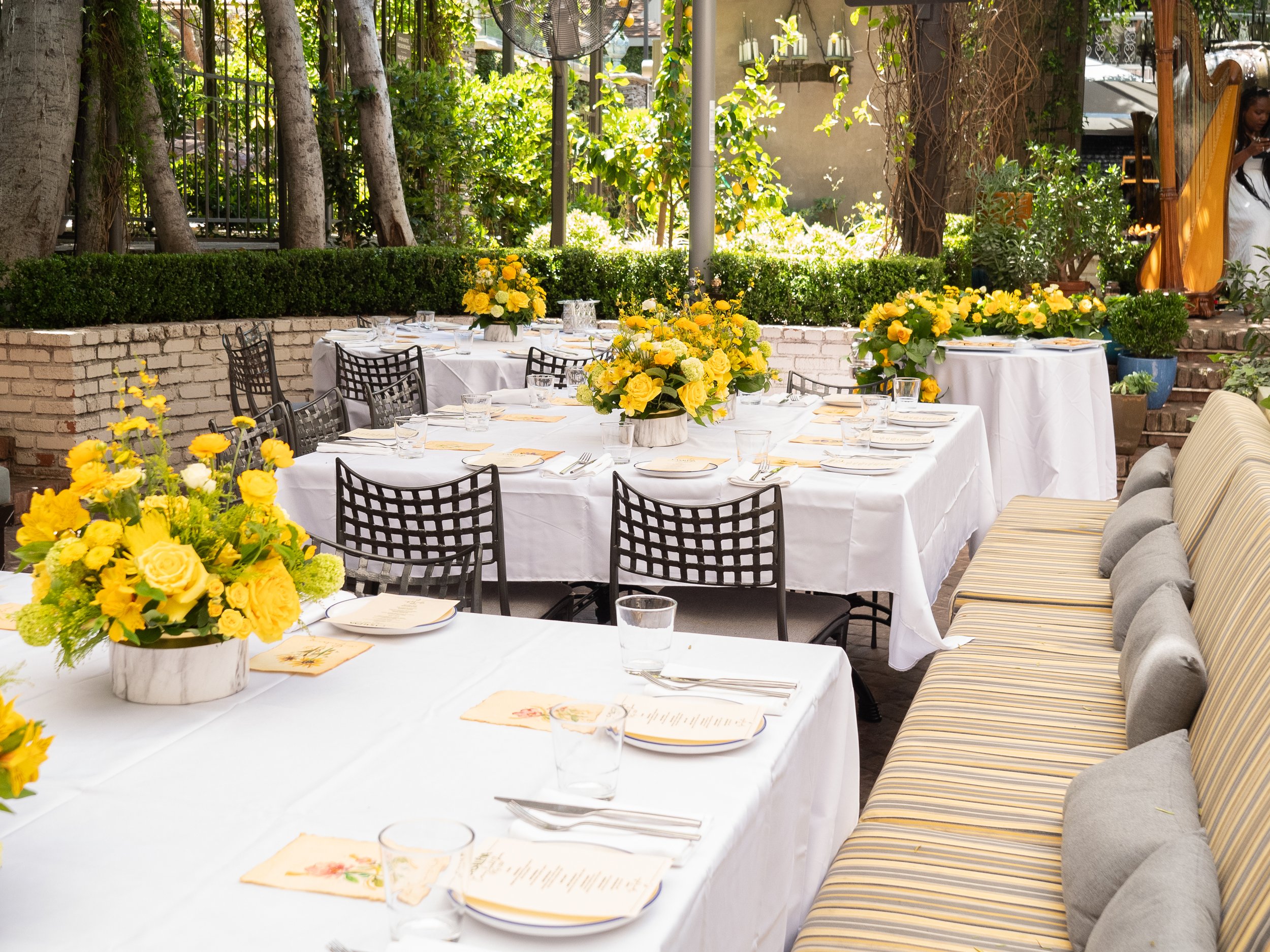 An outdoor dining area with tables covered in white tablecloths, decorated with yellow floral arrangements, and surrounded by chairs and cushioned benches. There are trees, bushes, and potted plants in the background, along with a brick wall, and a p