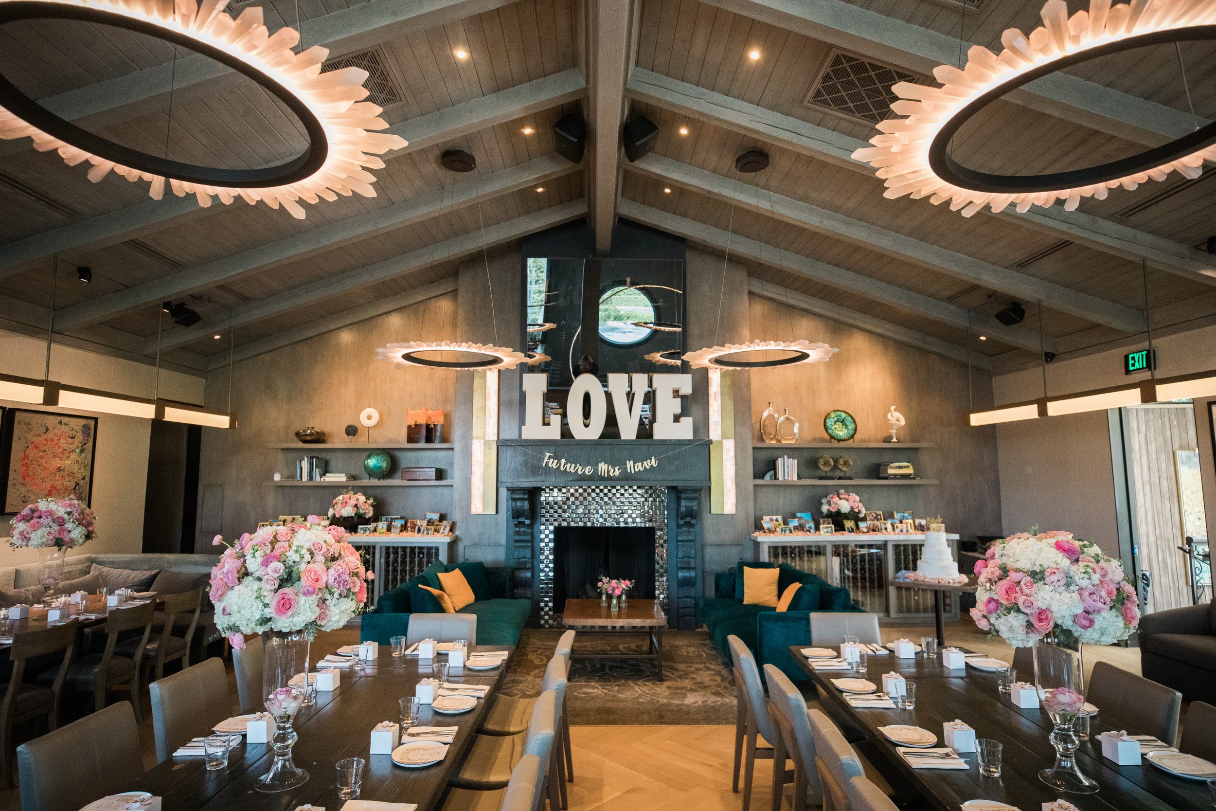 Elegant indoor event space decorated for a wedding, featuring large pink and white floral arrangements, tables set with white napkins and clear glasses, colorful cushions on dark green seating, a large 'LOVE' sign above the fireplace, and modern hang