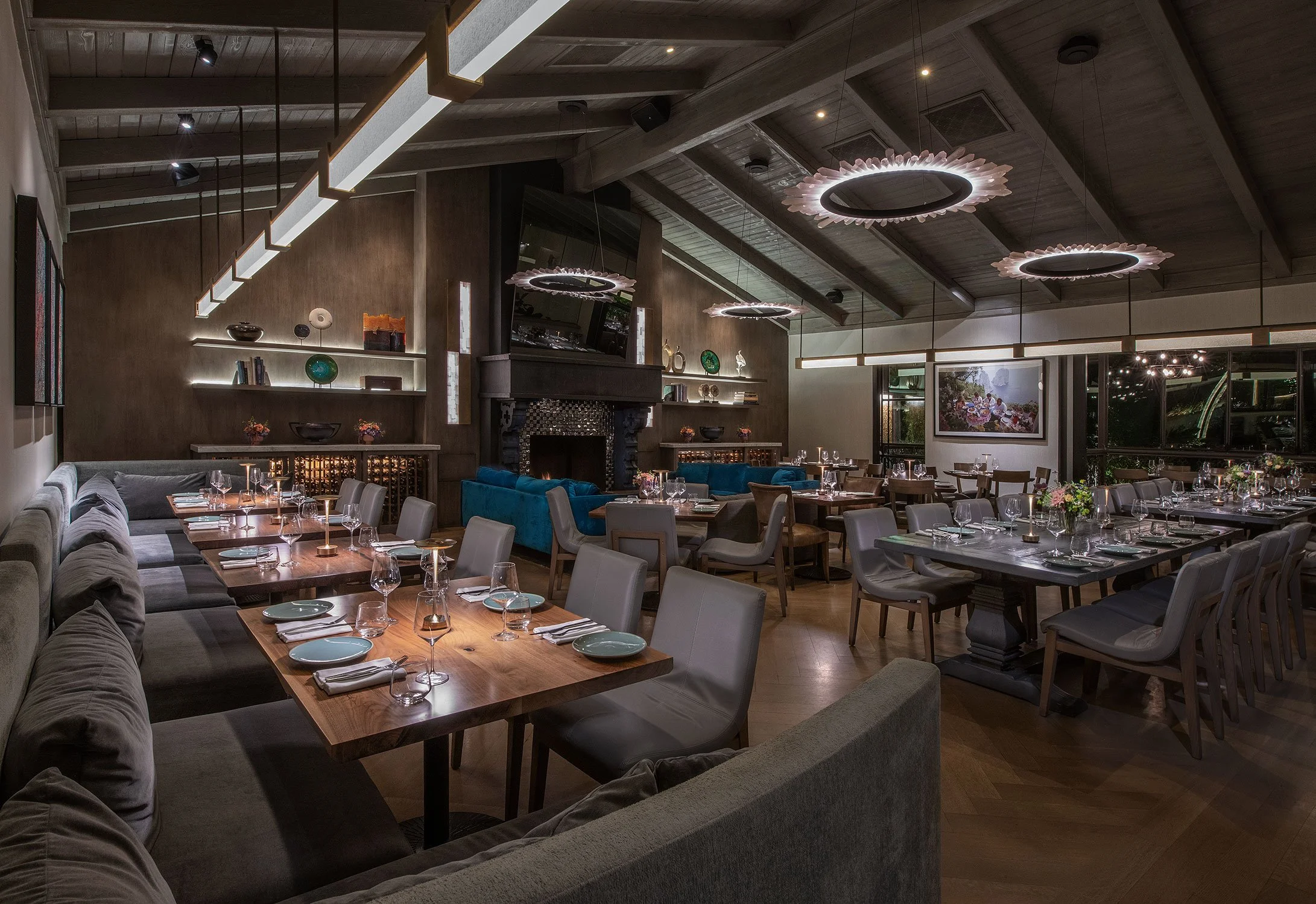 Interior view of a modern restaurant dining area with wood-paneled ceiling, stylish circular hanging light fixtures, and neatly set tables with place settings and flower centerpieces.