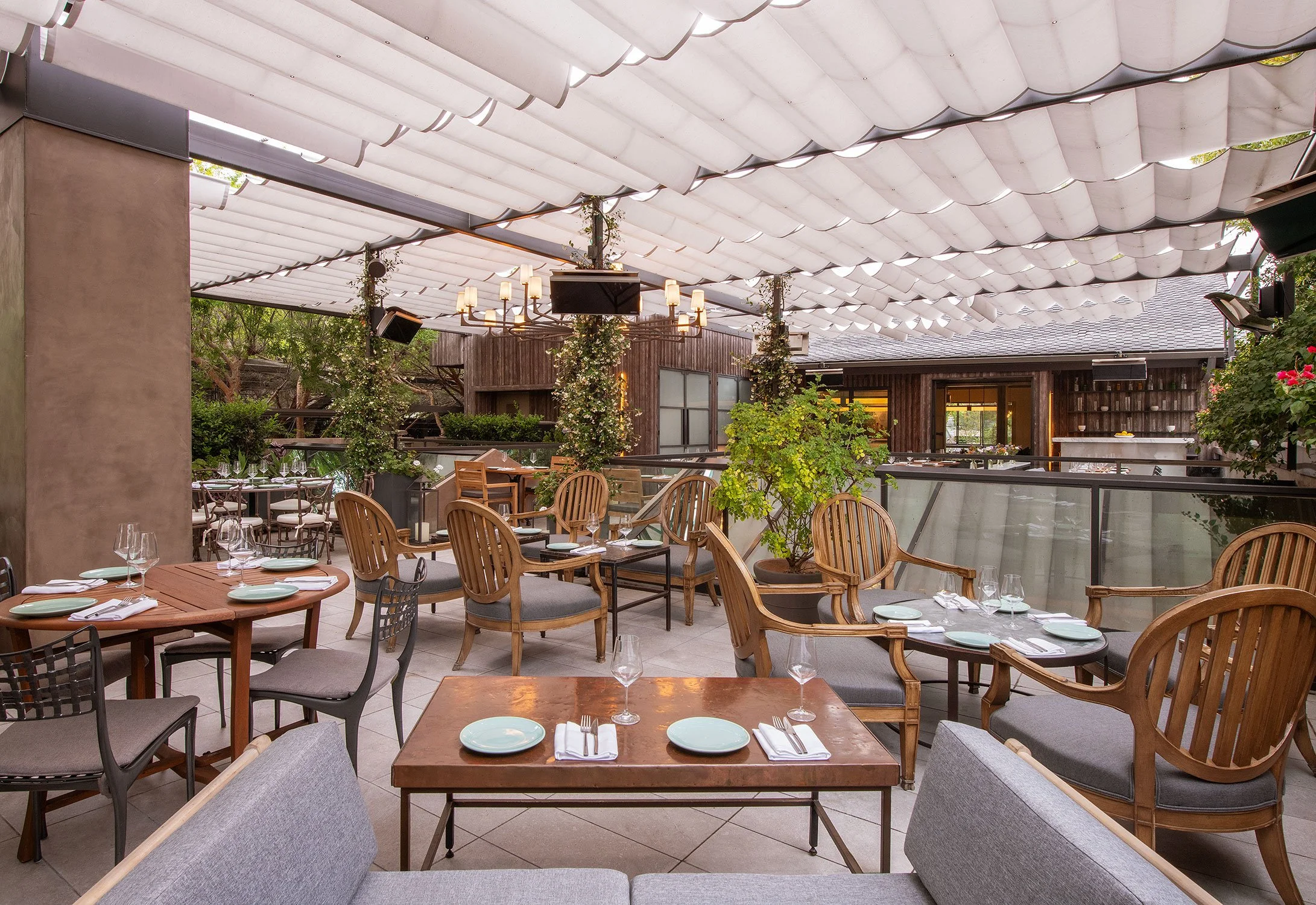 A restaurant patio with multiple dining tables set with plates, glasses, and napkins, surrounded by wooden and metal chairs, with umbrellas overhead and some greenery around.