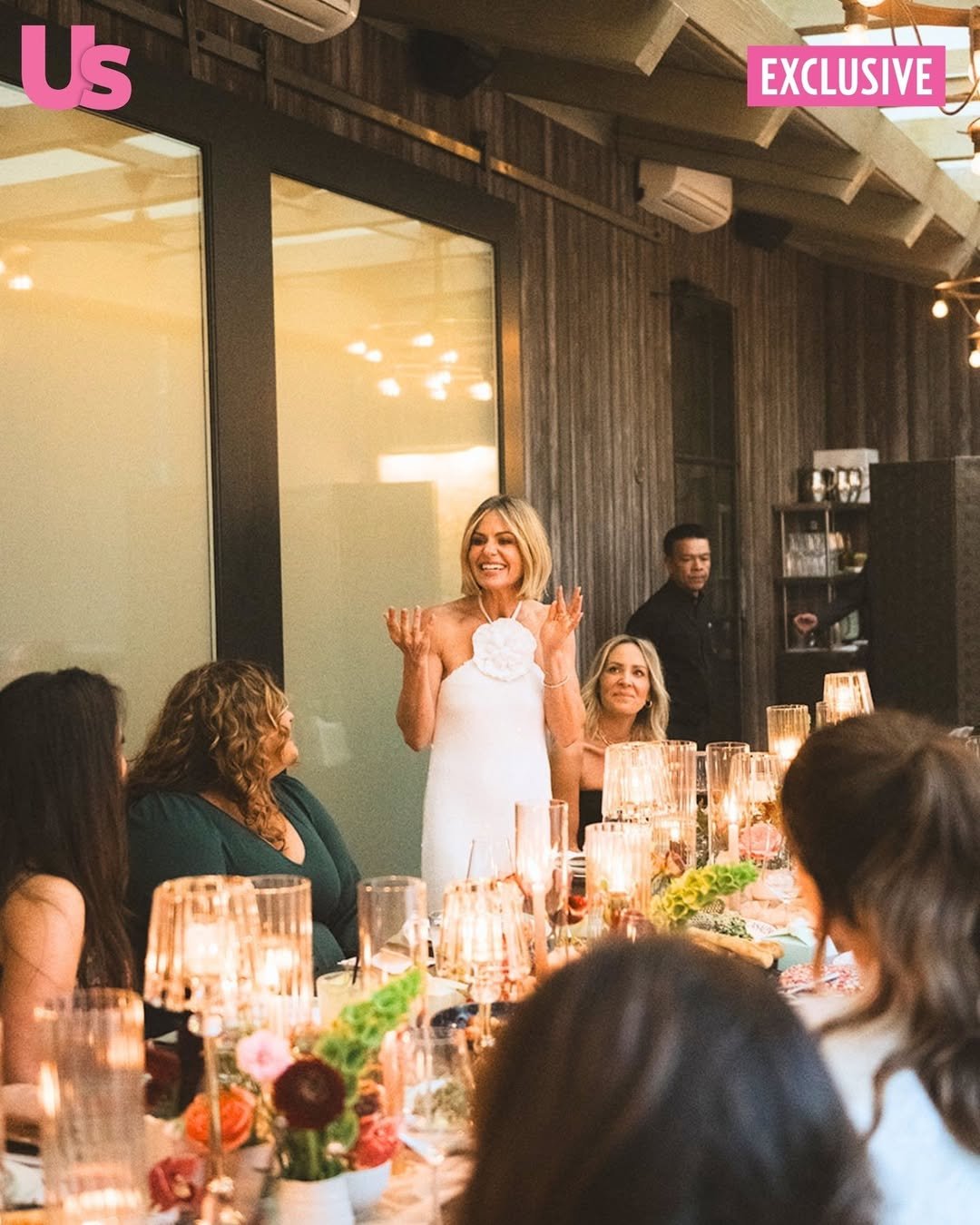 A woman in a white dress is standing and speaking to a group at a dinner table in a warmly lit restaurant with wood-paneled walls. Several women are seated around the table, listening to her, with candles and floral centerpieces on the table.