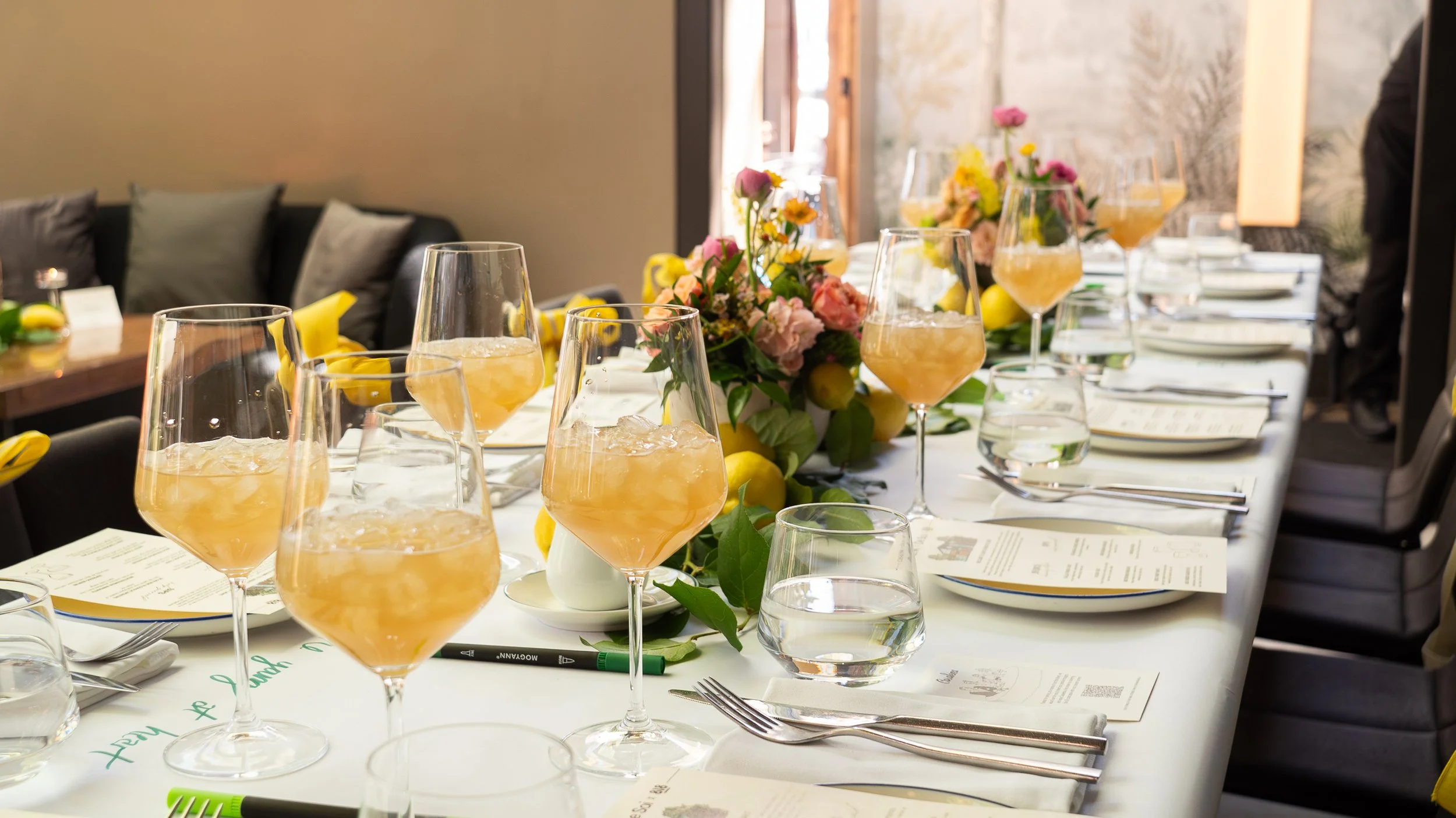 Table set for a celebration with glasses of drinks, floral centerpieces, place settings, and menus.