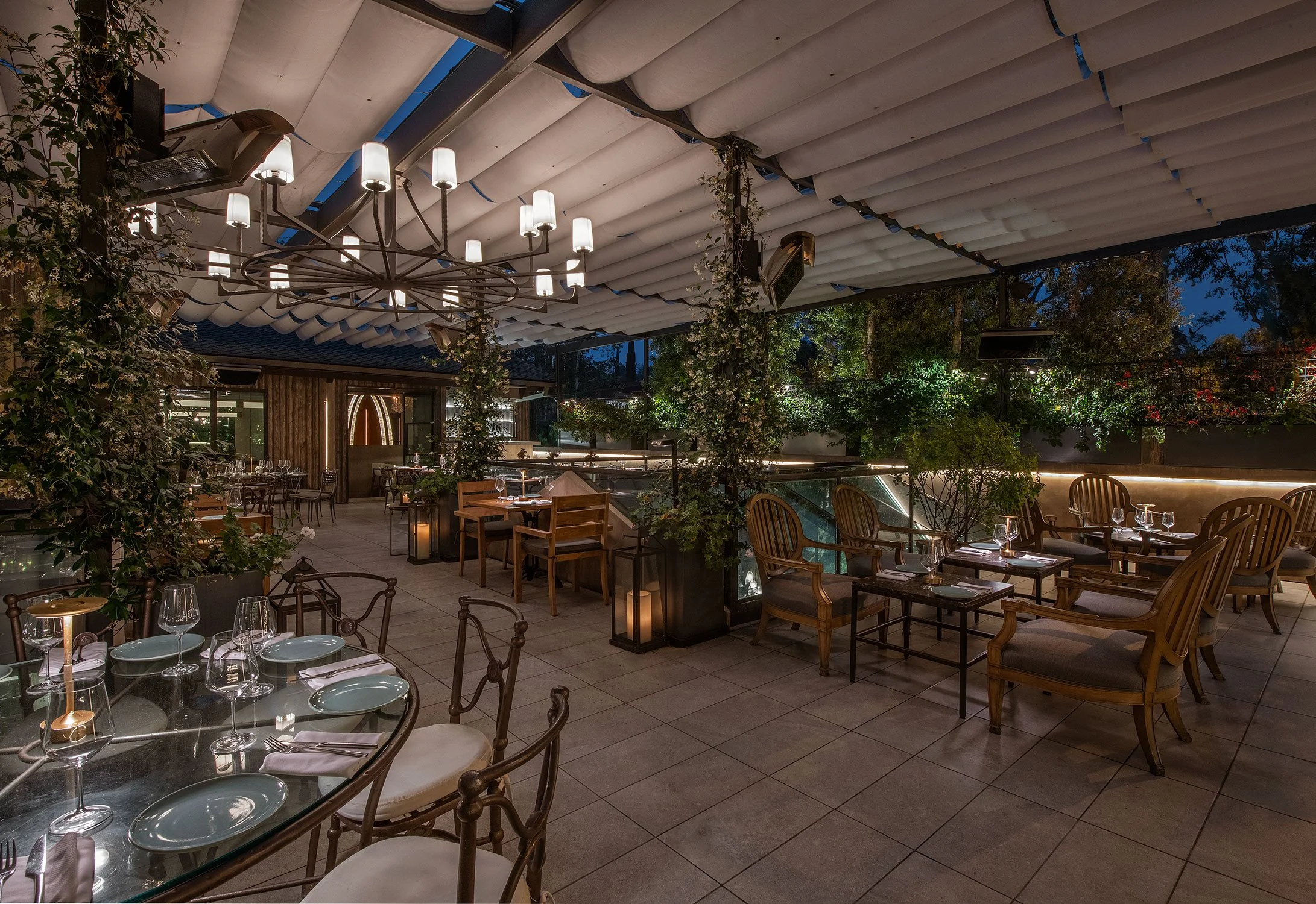 Indoor restaurant with glass-top tables, wooden chairs, and a ceiling with unique design elements, including cloud-like structures and hanging lights, surrounded by greenery and trees outside.