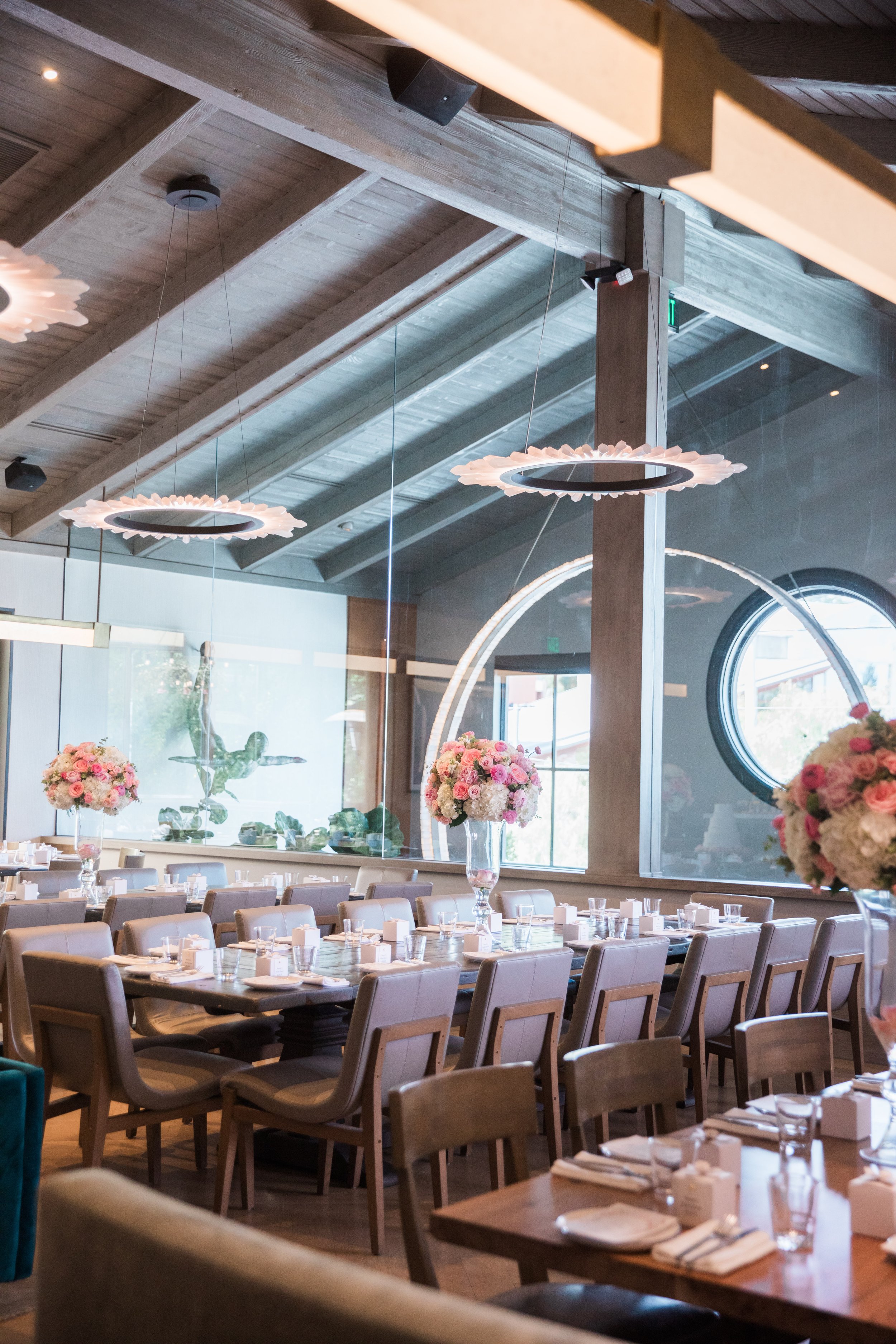 Interior of a modern restaurant decorated with floral centerpieces on tables, featuring large round paper flower ceiling lights and wooden beams.