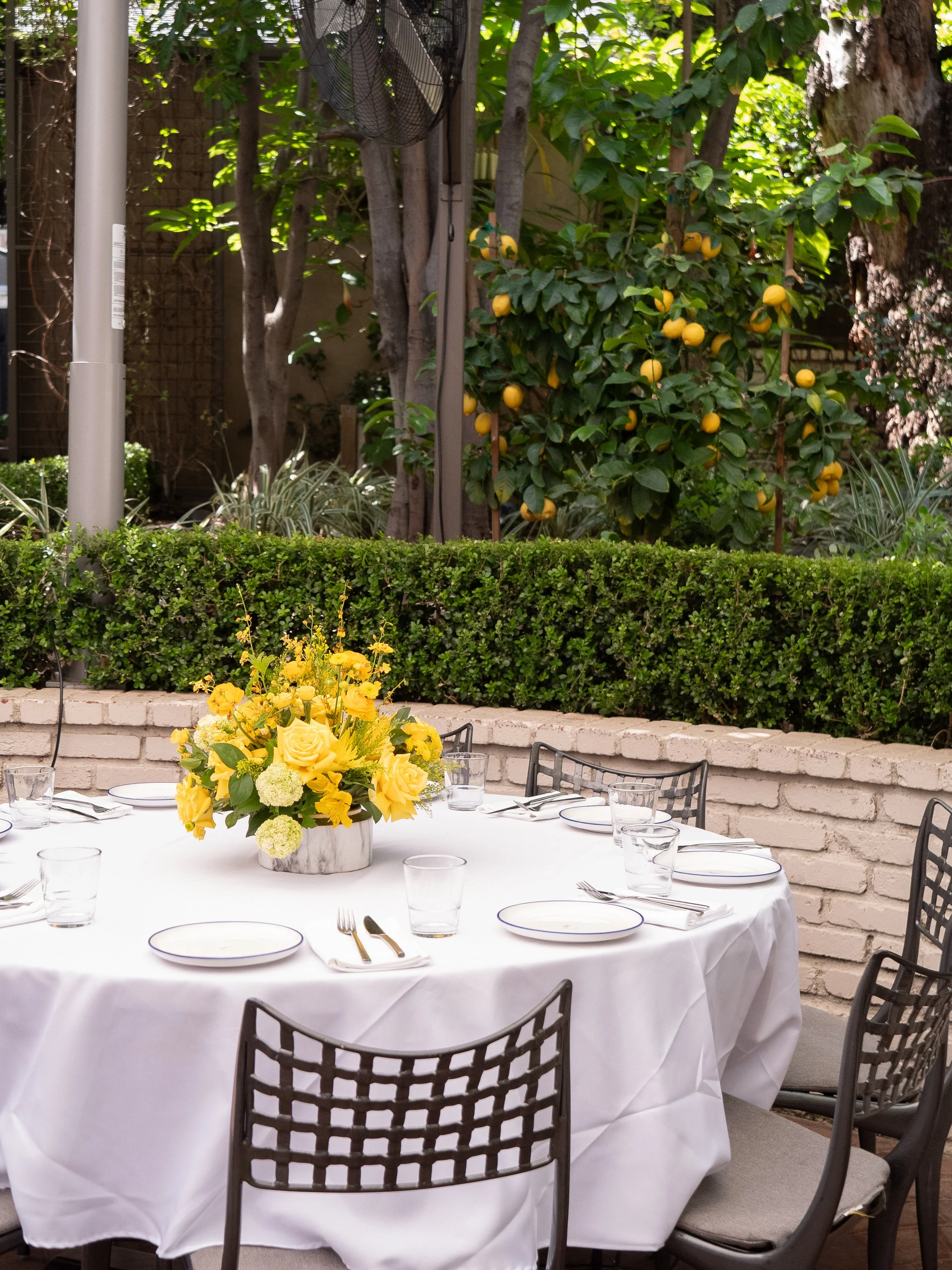 Round dining table covered with a white cloth, set for a meal with white plates, silverware, and drinking glasses. A yellow floral centerpiece decorates the table. The outdoor space is surrounded by a brick wall, green hedges, and trees, creating a l