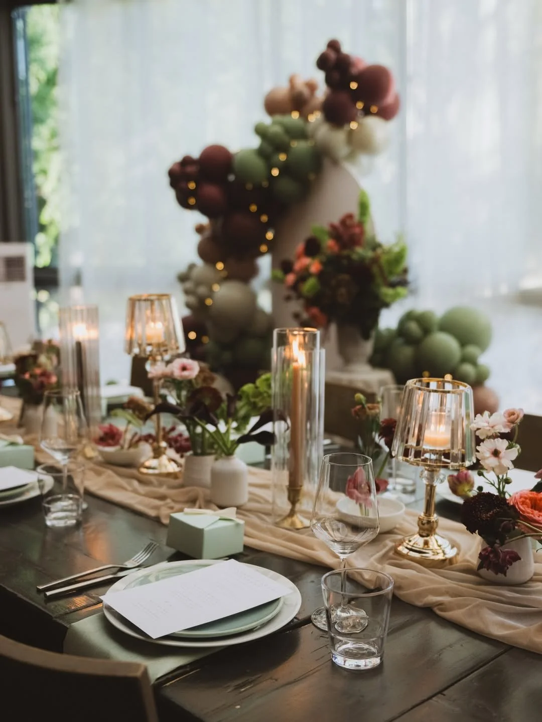 Elegant dining table decorated with flowers, candles, and a beige table runner, place settings with plates and glasses, and a blurred floral backdrop.