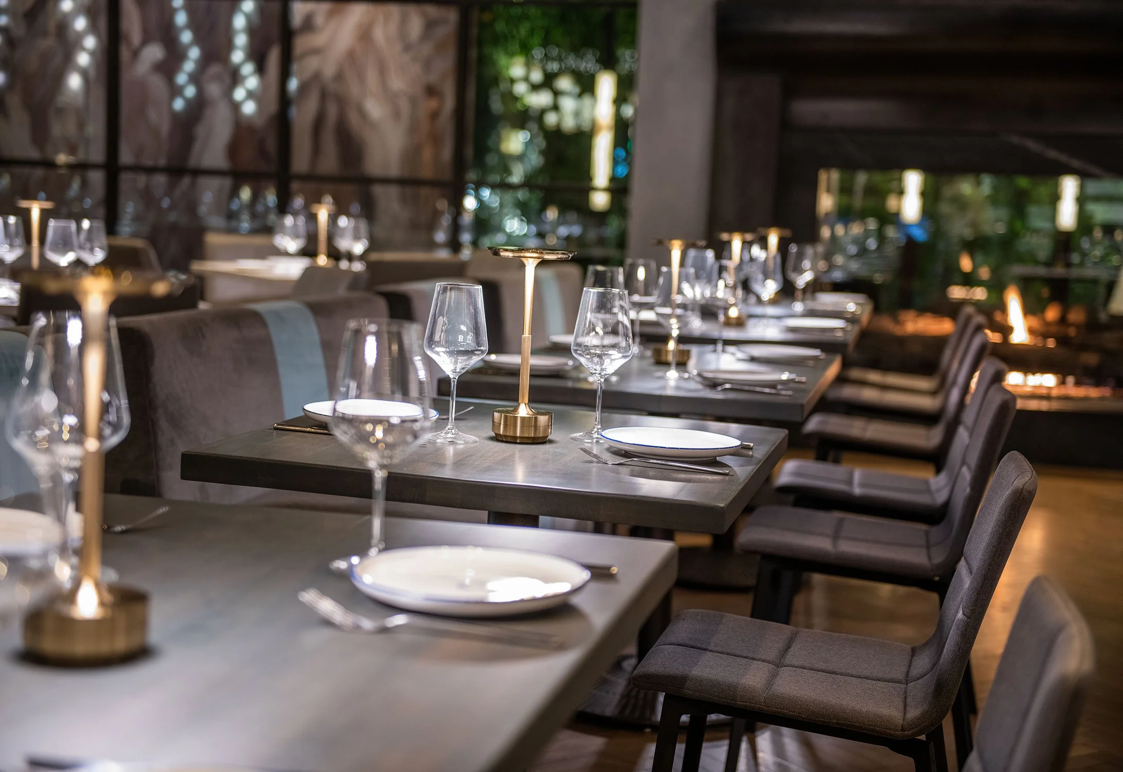 A restaurant dining area with multiple tables set with wine glasses, plates, and silverware. The room features modern decor with dim lighting and dark furniture, with large windows and greenery visible outside.