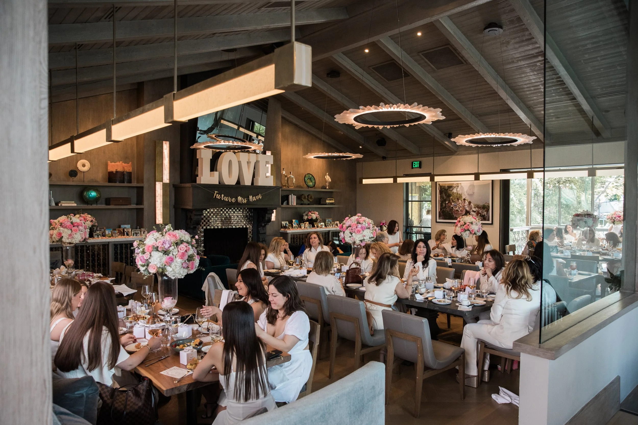 A wedding reception with women sitting at tables decorated with pink flowers, enjoying meal and conversation, inside a modern venue with large windows and decorative lighting.