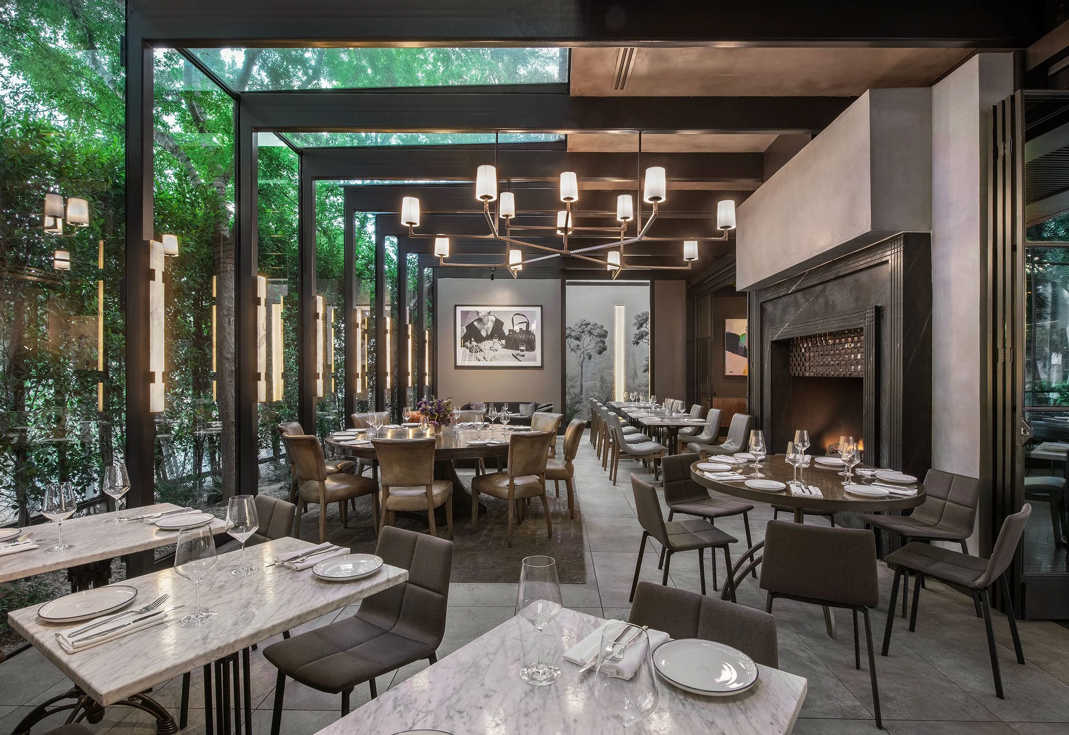 Elegant restaurant interior with large windows, greenery outside, modern chandelier, and neatly set tables with glassware and plates.