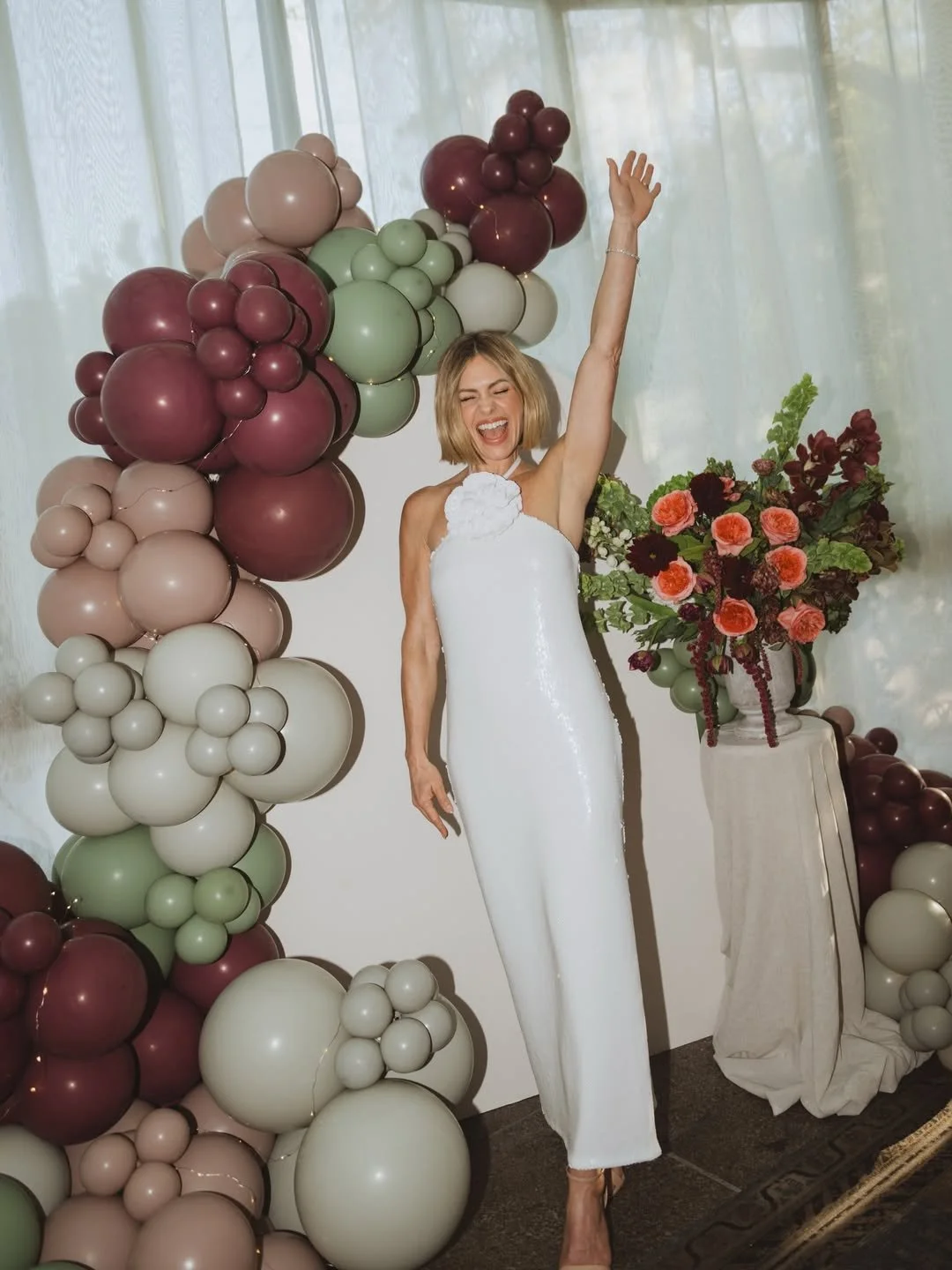 Woman in a white dress celebrating with balloons and flowers.