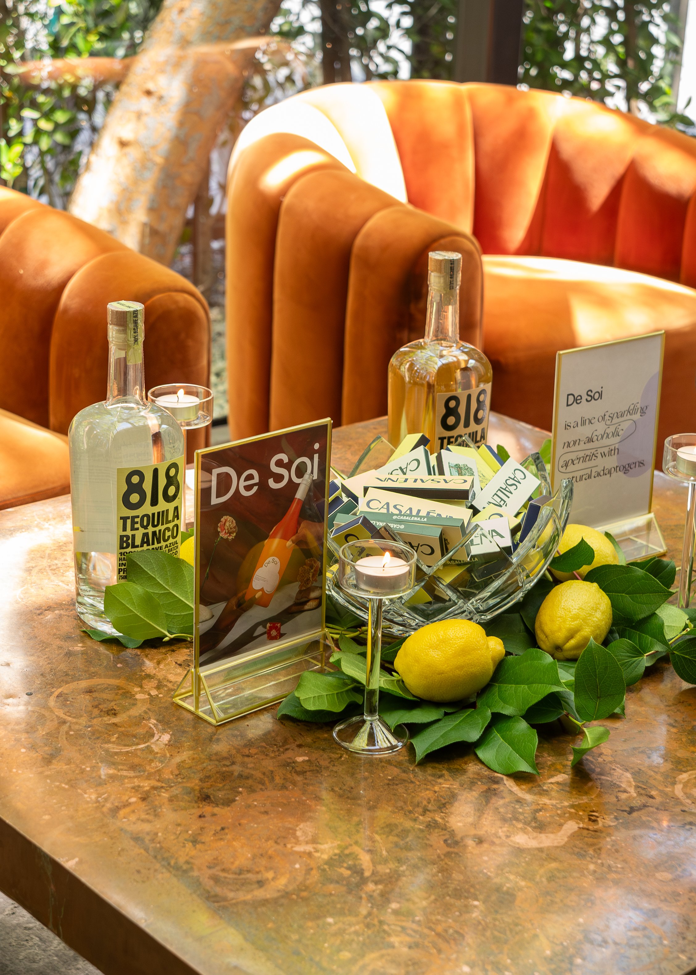 A table with bottles of tequila, lemons, candles, and promotional signs for De Soi and 818 Tequila Blanco. Green leaves and a bowl of lemons decorate the table, with chairs and a window with greenery in the background.