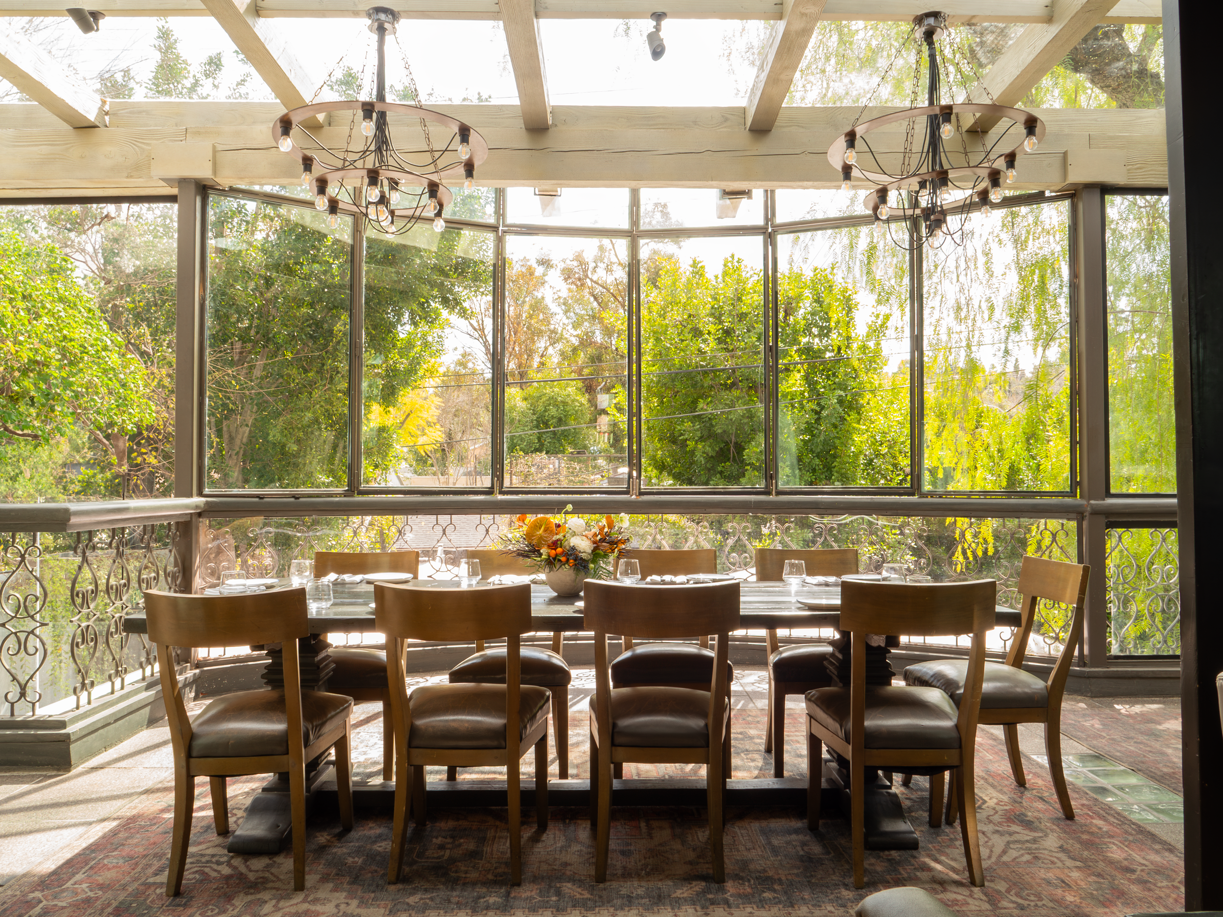 A dining area with a large wooden table surrounded by chairs, set for a meal, with a floral centerpiece. Large windows behind the table reveal an outdoor view of trees and blue sky, with a wooden ceiling overhead and two rustic chandeliers hanging fr