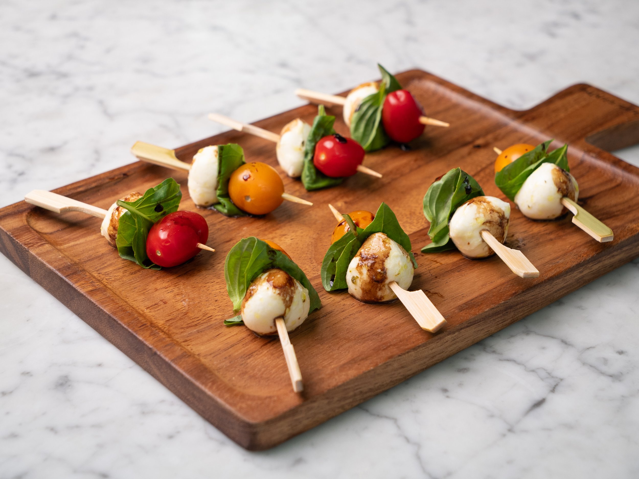 Skewers of mozzarella balls, cherry tomatoes, basil leaves, drizzled with balsamic glaze on a wooden tray.