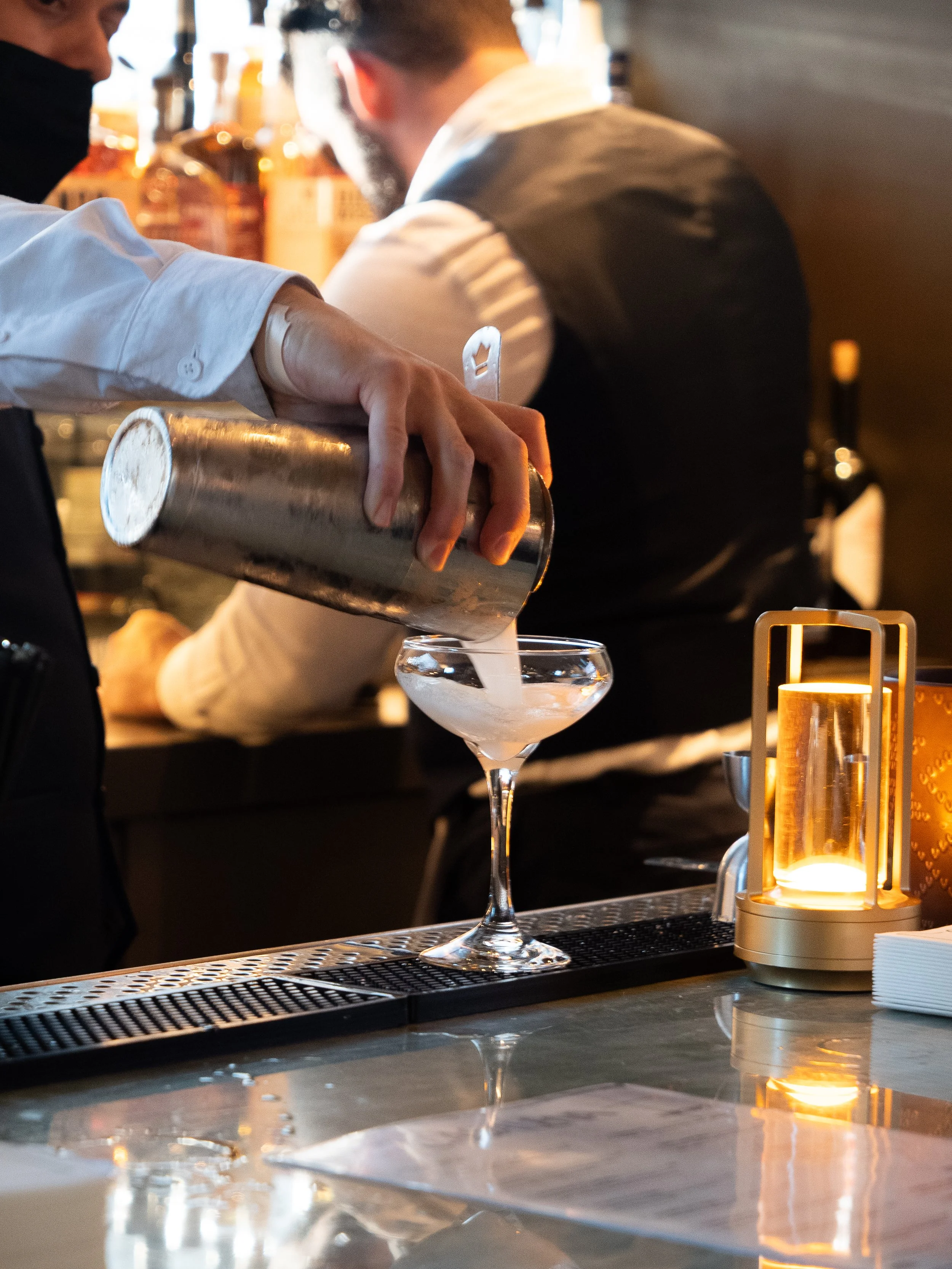 Bartender pouring a cocktail into a glass at a bar counter with candles and liquor bottles in the background.