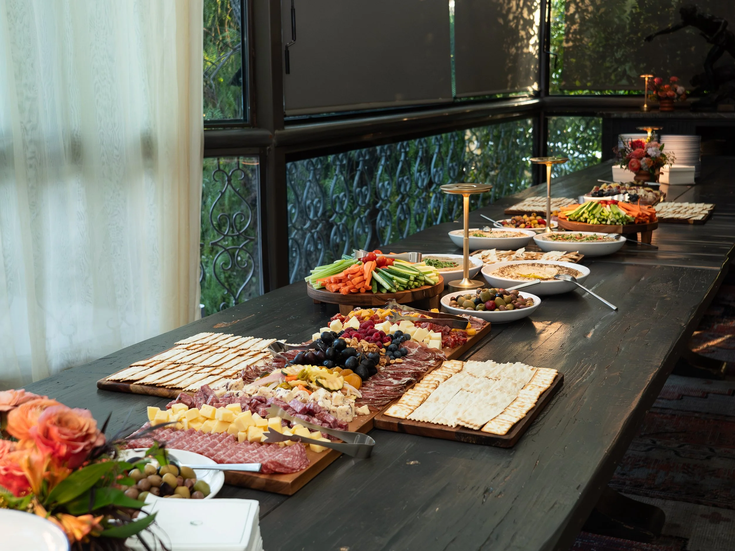 A table filled with various cheeses, sliced meats, fresh vegetables, grapes, and crackers, set up for a buffet with a window showing outdoor greenery in the background.