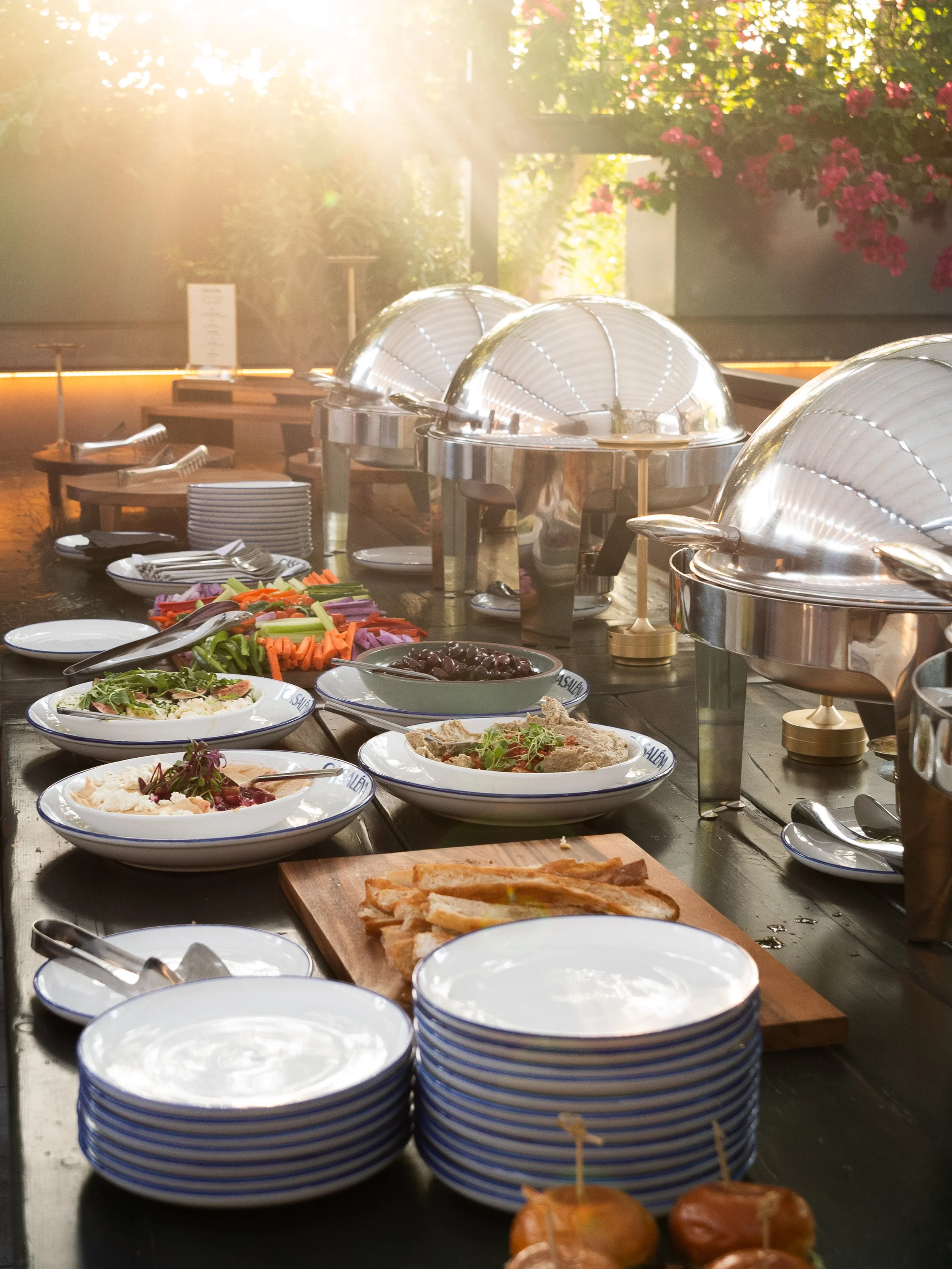 A buffet table with chafing dishes, plates, and bowls filled with salads, vegetables, and appetizers, set in a well-lit area with sunlight streaming in and surrounding greenery.