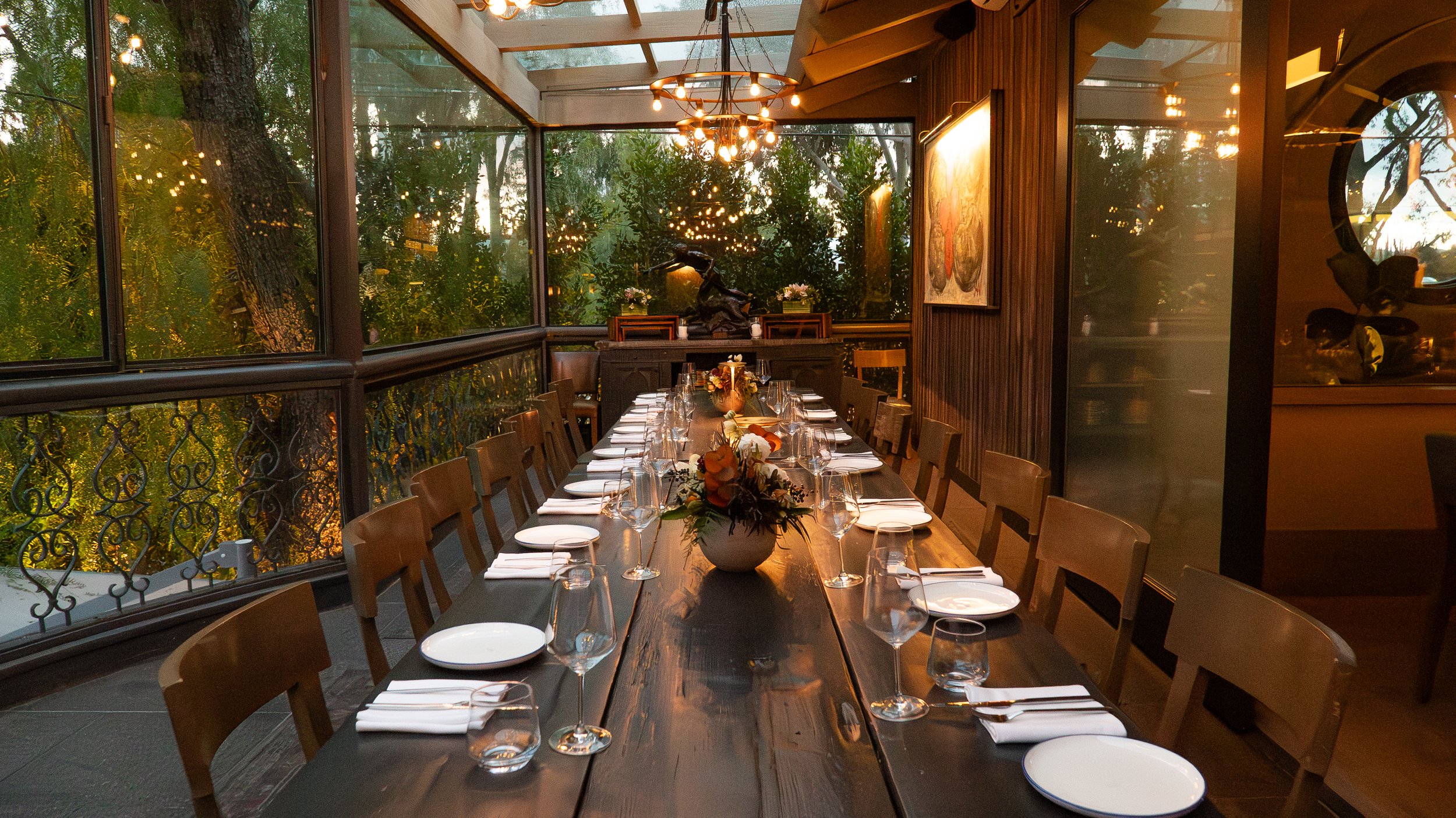 A long dining table set for a meal with glasses, plates, and a floral centerpiece inside a glass-walled restaurant with trees visible outside.