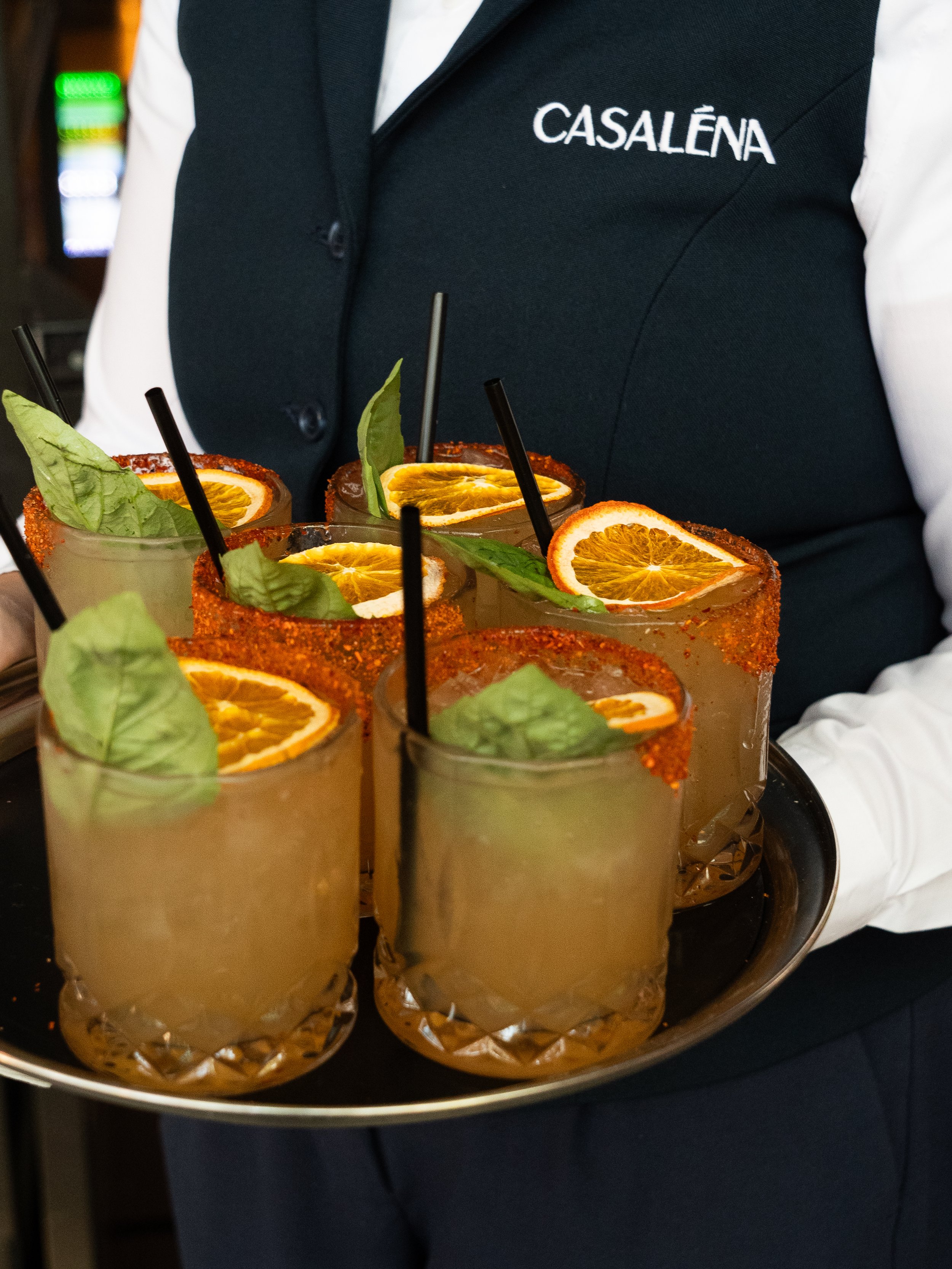 A person wearing a black vest with the name 'CASALÉNA' embroidered on it, holding a tray with multiple margarita glasses garnished with orange slices and basil leaves.