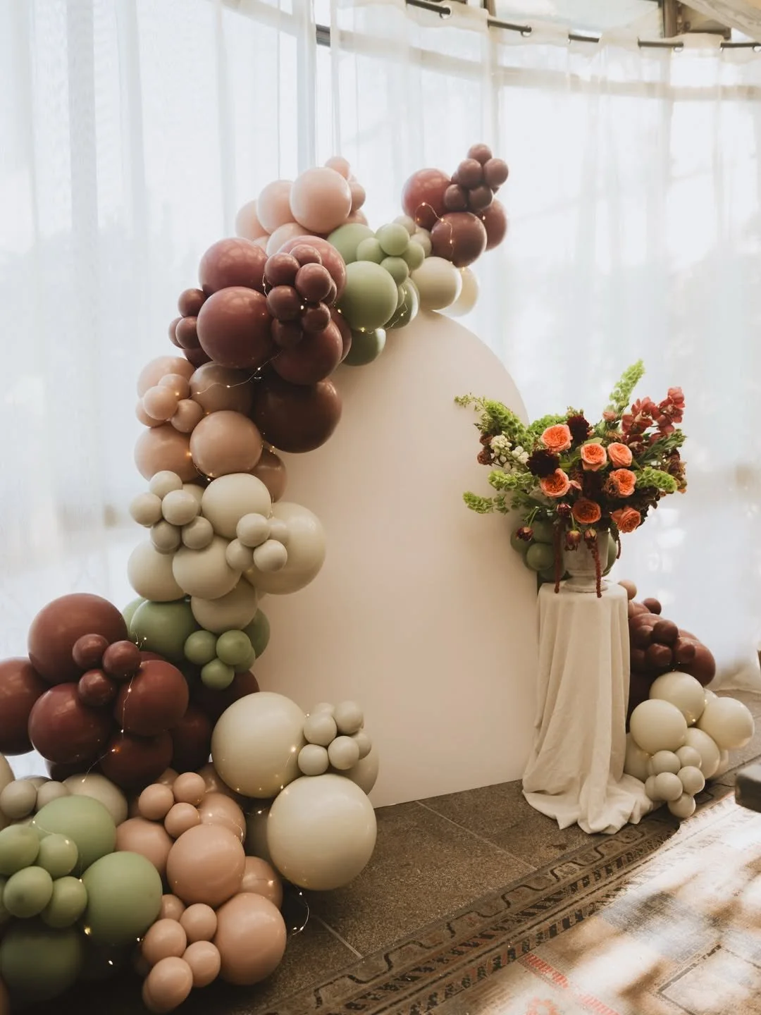 Decorative balloon arch with a white backdrop and a floral arrangement on a small table.