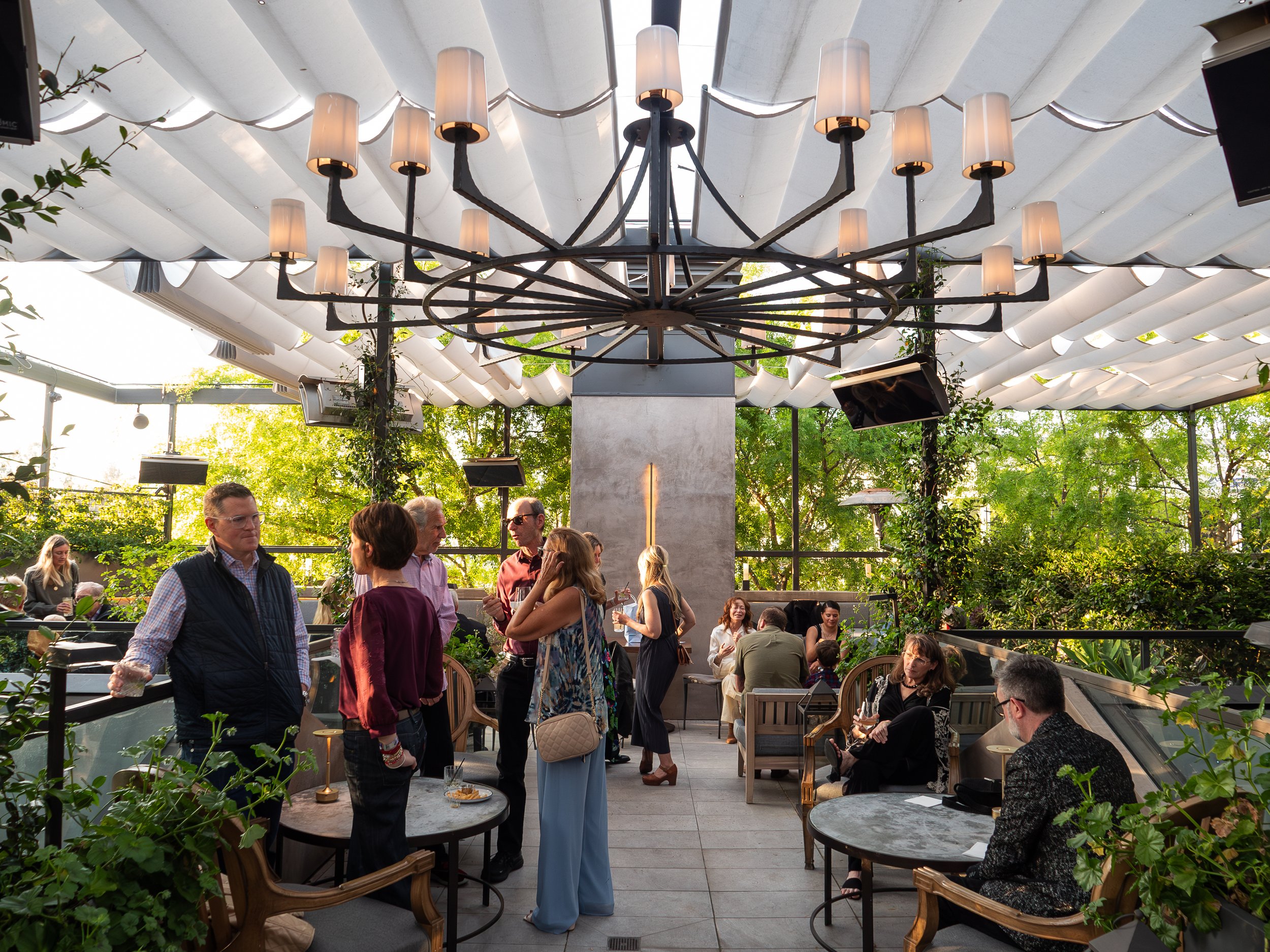 People socializing inside an indoor-outdoor restaurant or cafe with a large decorative chandelier and greenery around.
