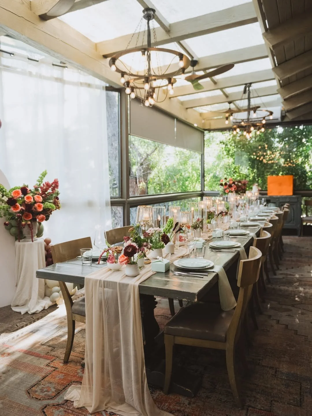 Elegant indoor dining setup with a long table, floral centerpieces, candles, glassware, and tableware, decorated for a special event.
