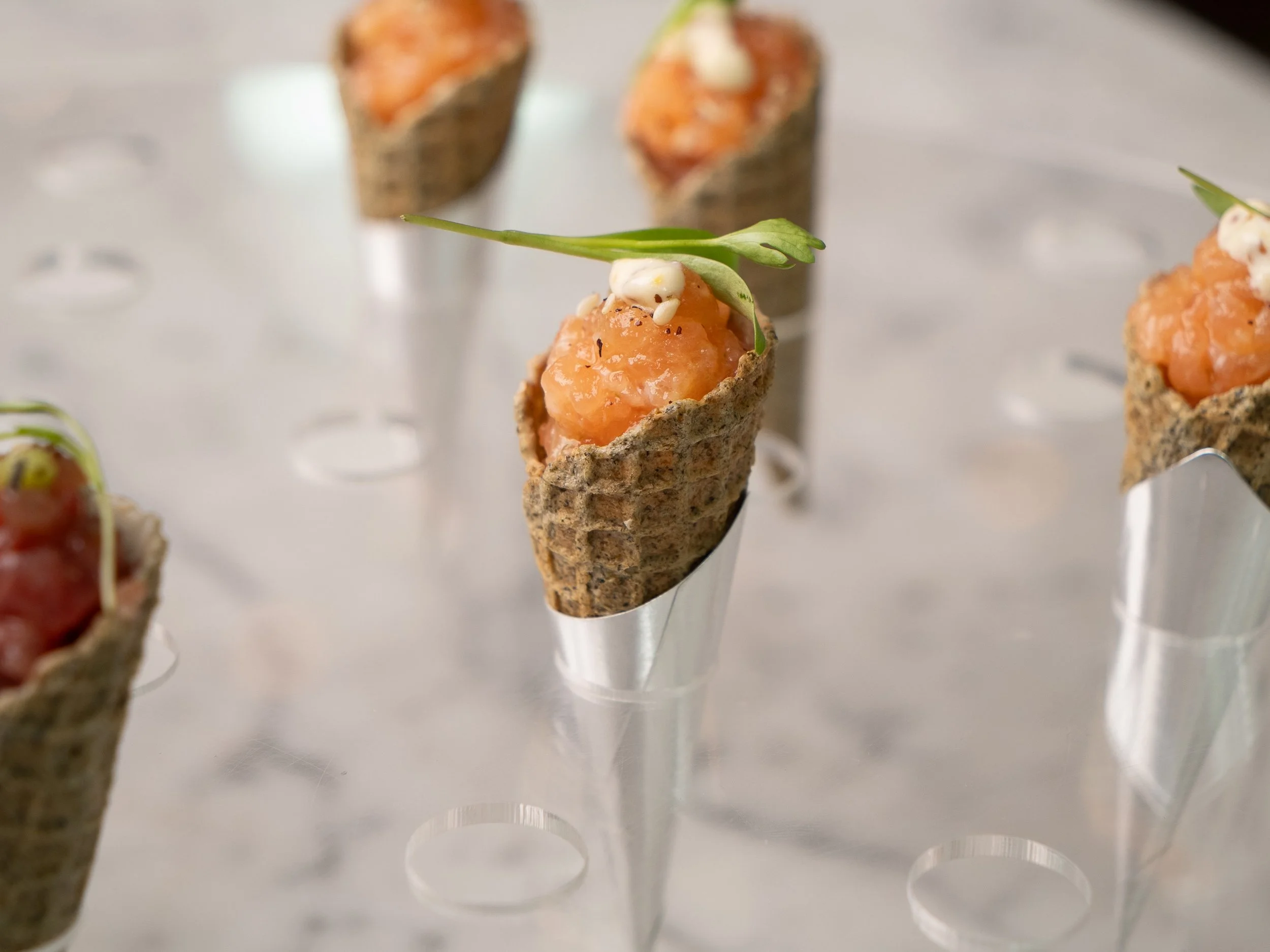 Close-up of smoked salmon bites in edible cups garnished with microgreens on a metal display stand.