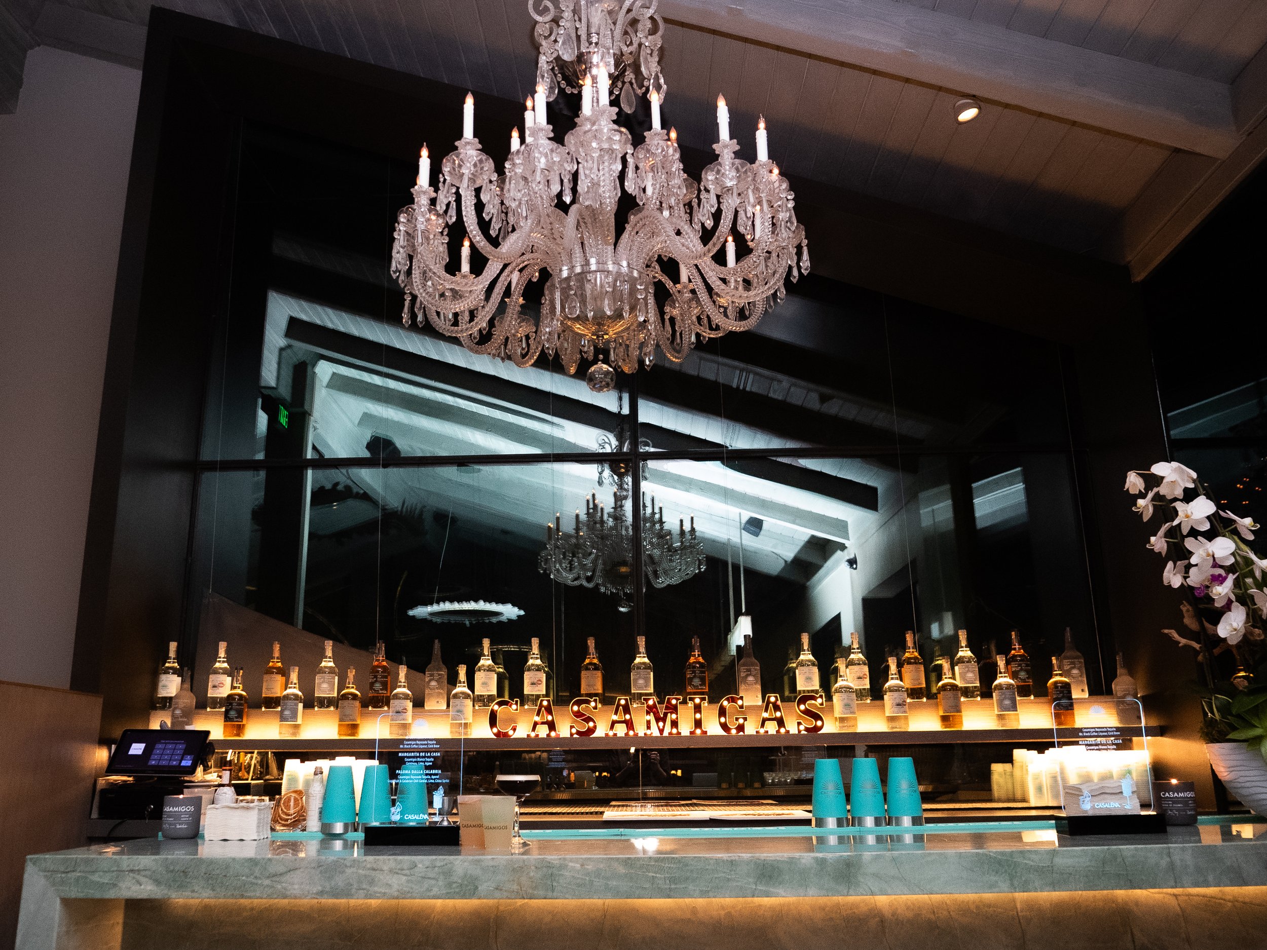 An elegant crystal chandelier hanging from a wooden ceiling above a bar counter. The bar features bottles of alcohol lined up with warm lighting, a menu, and turquoise-colored cups. There are floral decorations on the right side and a reflective mirr