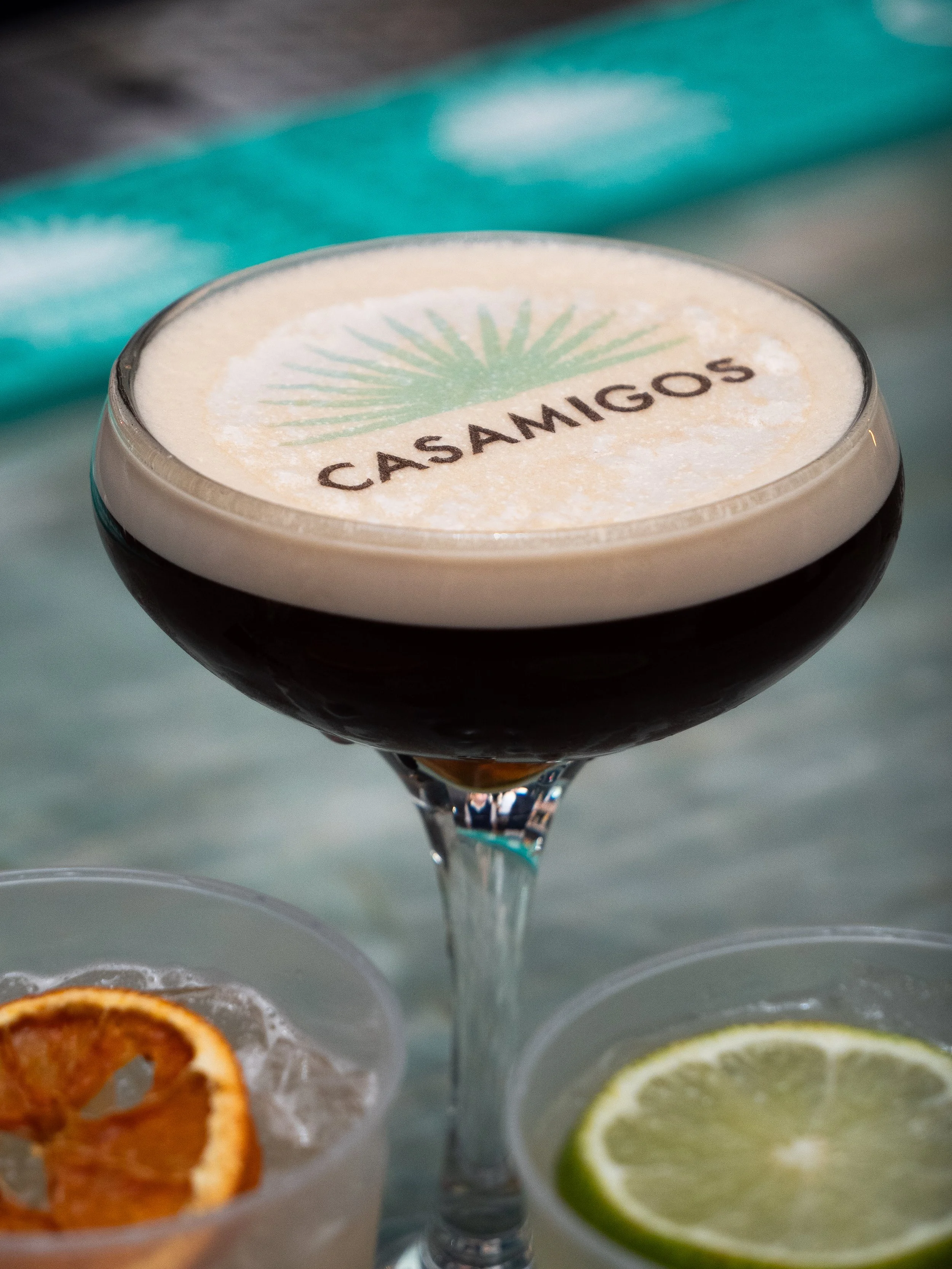 A cocktail glass with foam on top displaying the Casamigos logo, with lemon and lime slices and dried orange garnishes nearby.