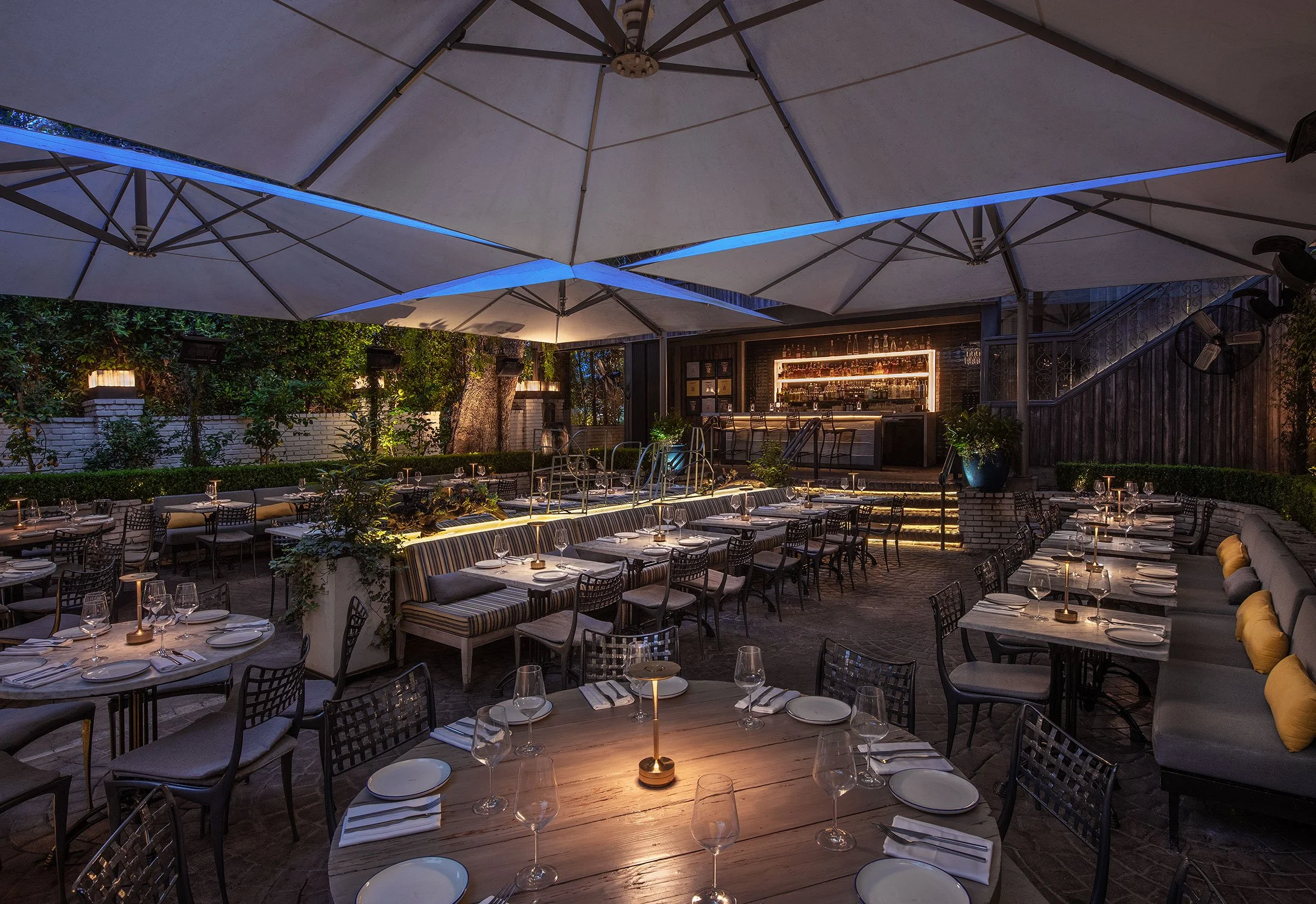 An outdoor dining area at night with tables set with glasses, plates, and napkins, illuminated by candles and ambient lighting, covered by large umbrellas, and surrounded by greenery and a bar area in the background.