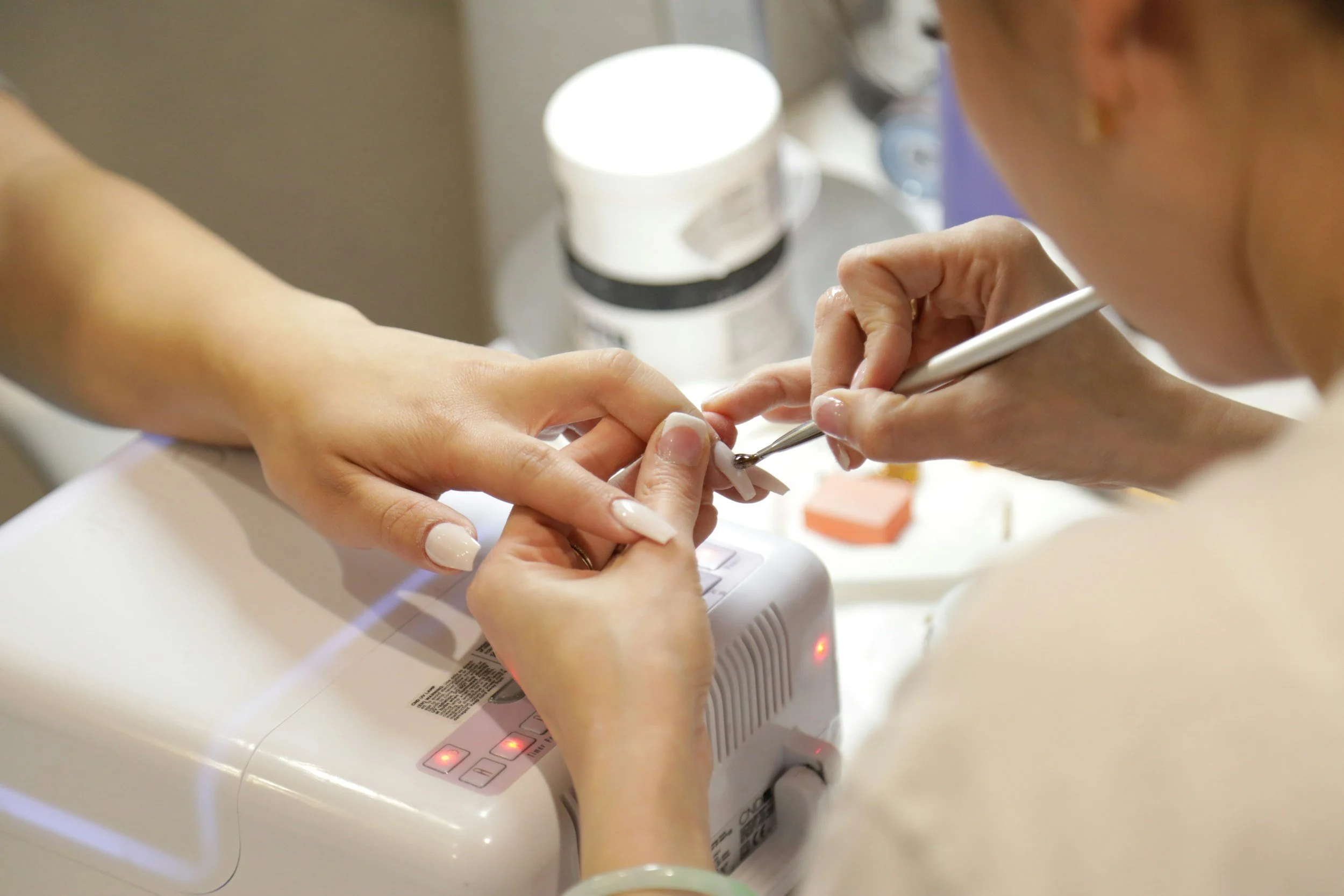 Close-up of a manicure in progress with a person applying nail polish to a client's nails using a brush, and a UV lamp on the table.