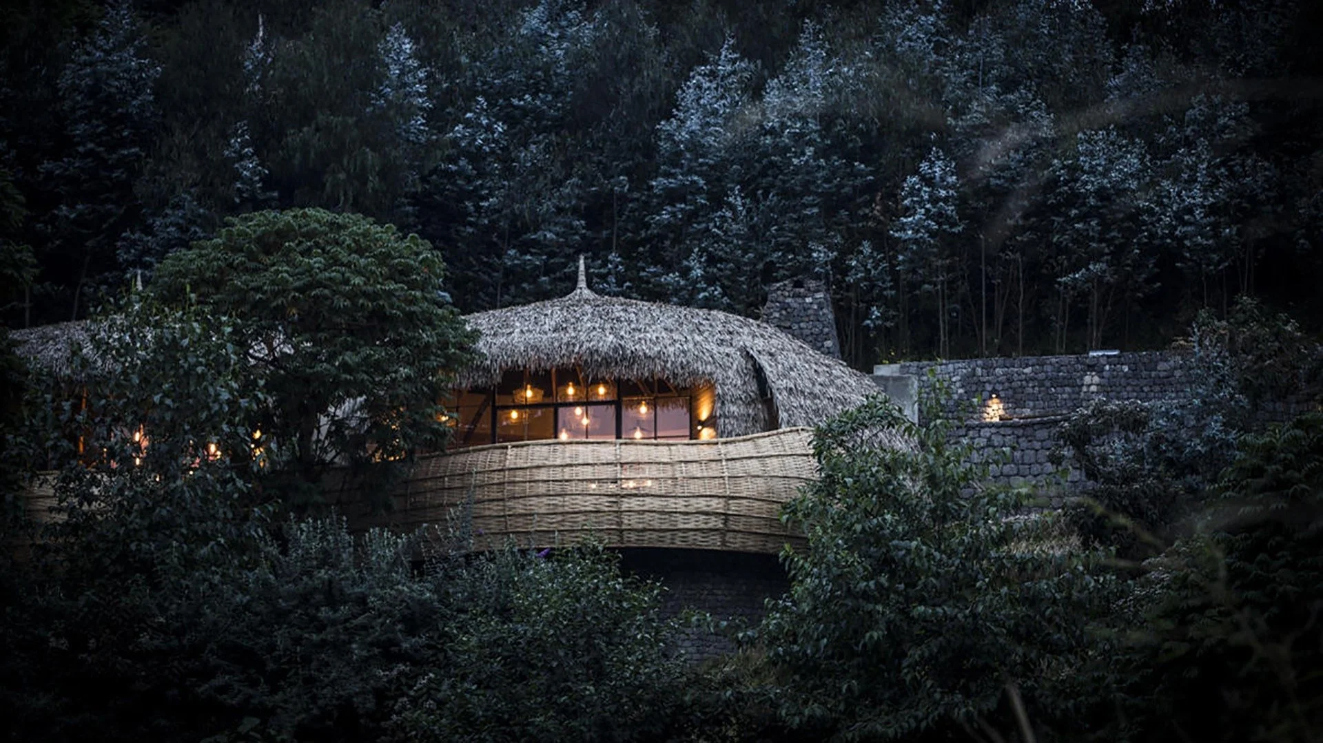 A cozy house with a thatched roof nestled among trees at night, illuminated by warm lights visible through large windows.