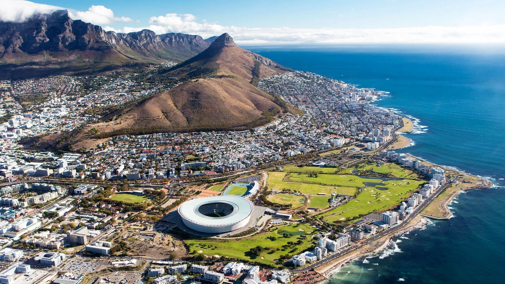Aerial view of Cape Town, South Africa, showing the city, mountains, and coastline with a stadium and green park area near the shore.