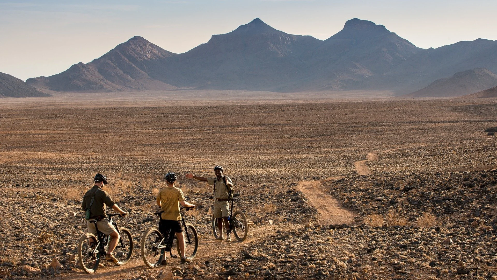 Three people in helmets and outdoor gear with bicycles standing on a dirt trail in a barren, rocky desert landscape with mountains in the distance.