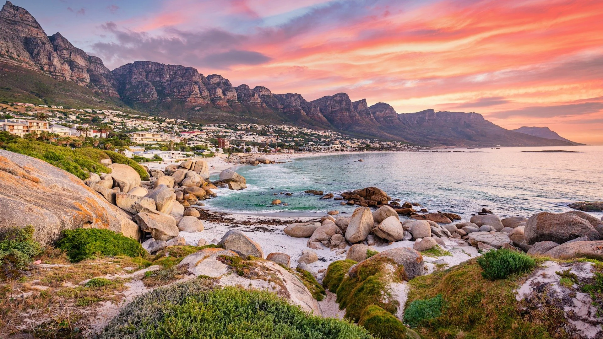 Scenic view of a rocky beach at sunset with colorful sky, mountains in the background, and a city along the shoreline.