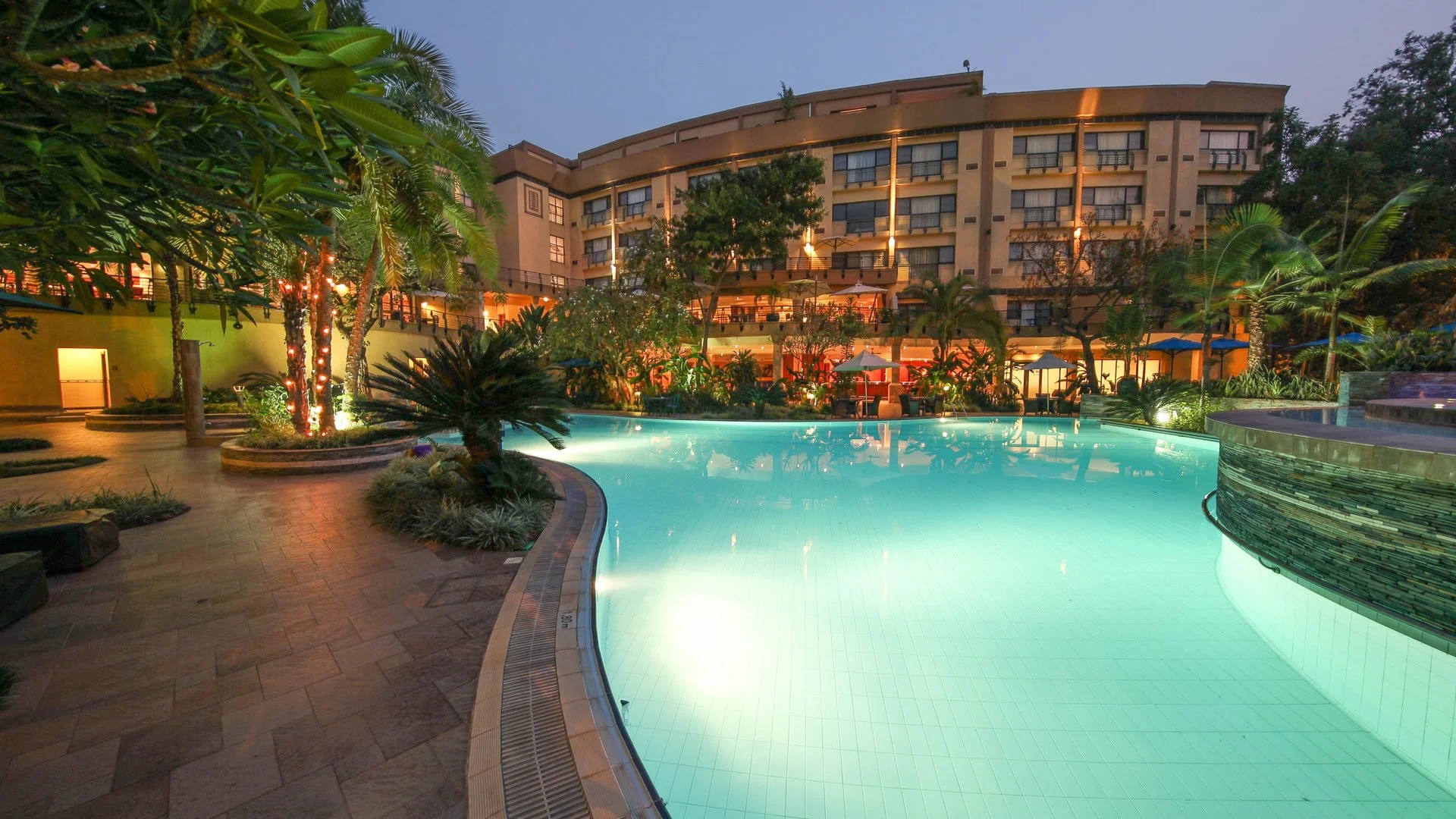 Illuminated swimming pool at sunset surrounded by tropical trees and a multi-story hotel with balconies.