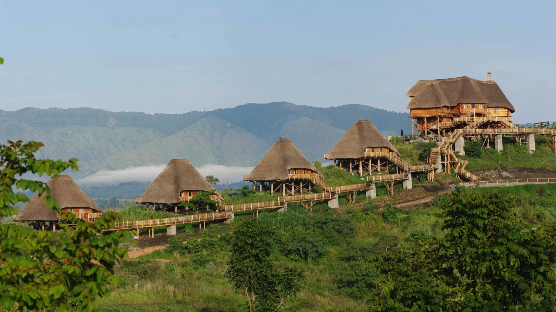 A series of traditional thatched-roof houses on stilts arranged on a hillside with mountains in the background and lush green trees in the foreground